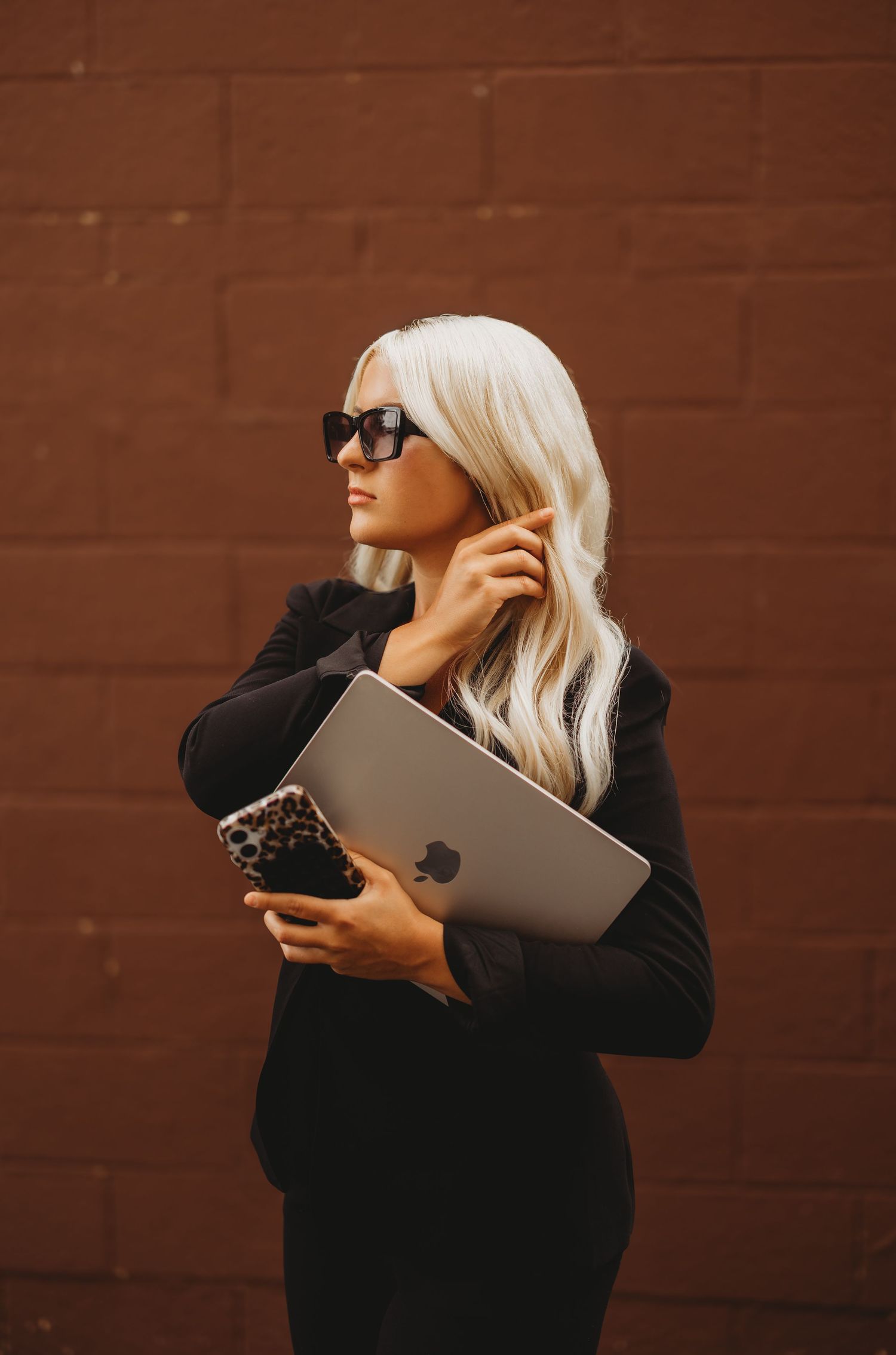 Blonde woman in black blazer and sunglasses holding MacBook and leopard phone against brick wall.
