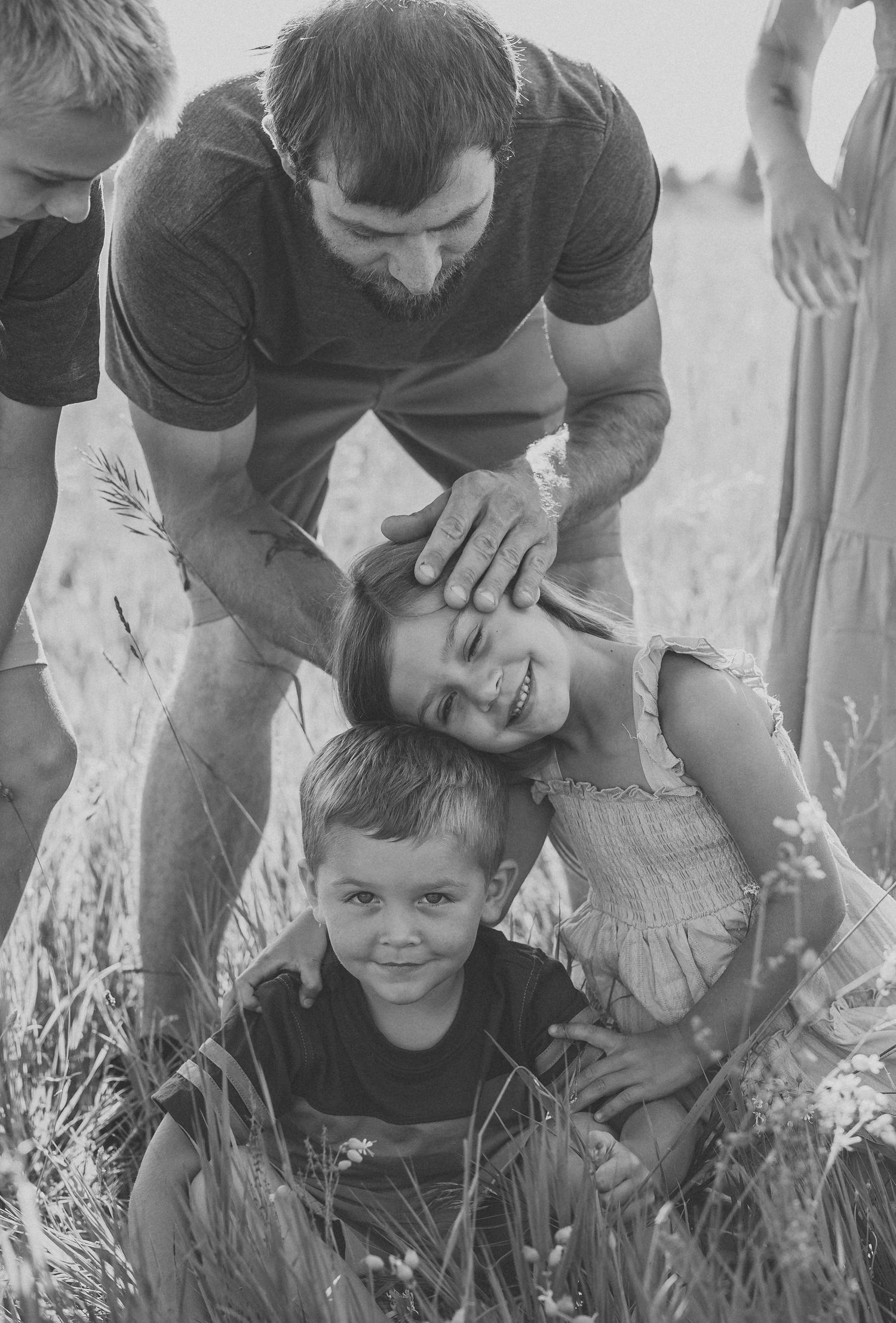 Black and white photo of a bearded father playfully ruffling his smiling daughter's hair while son poses in a field.