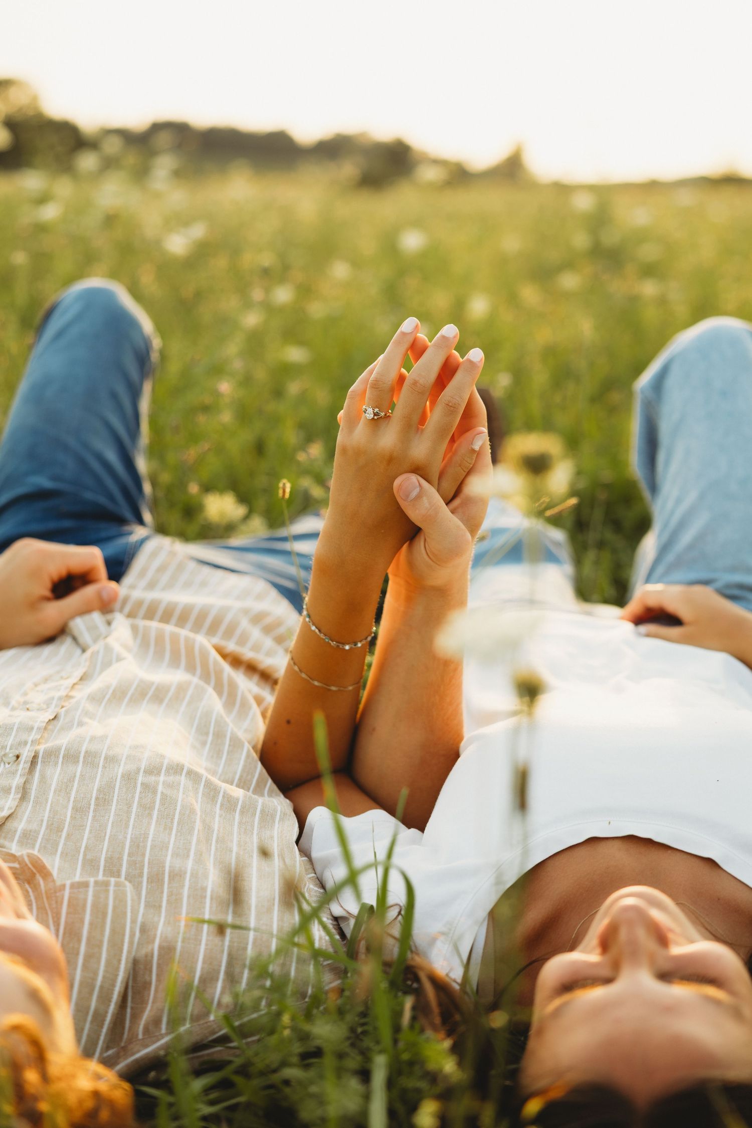 Couple holding hands while lying in a sunny wildflower meadow, wearing jeans and a striped dress with gold jewelry.