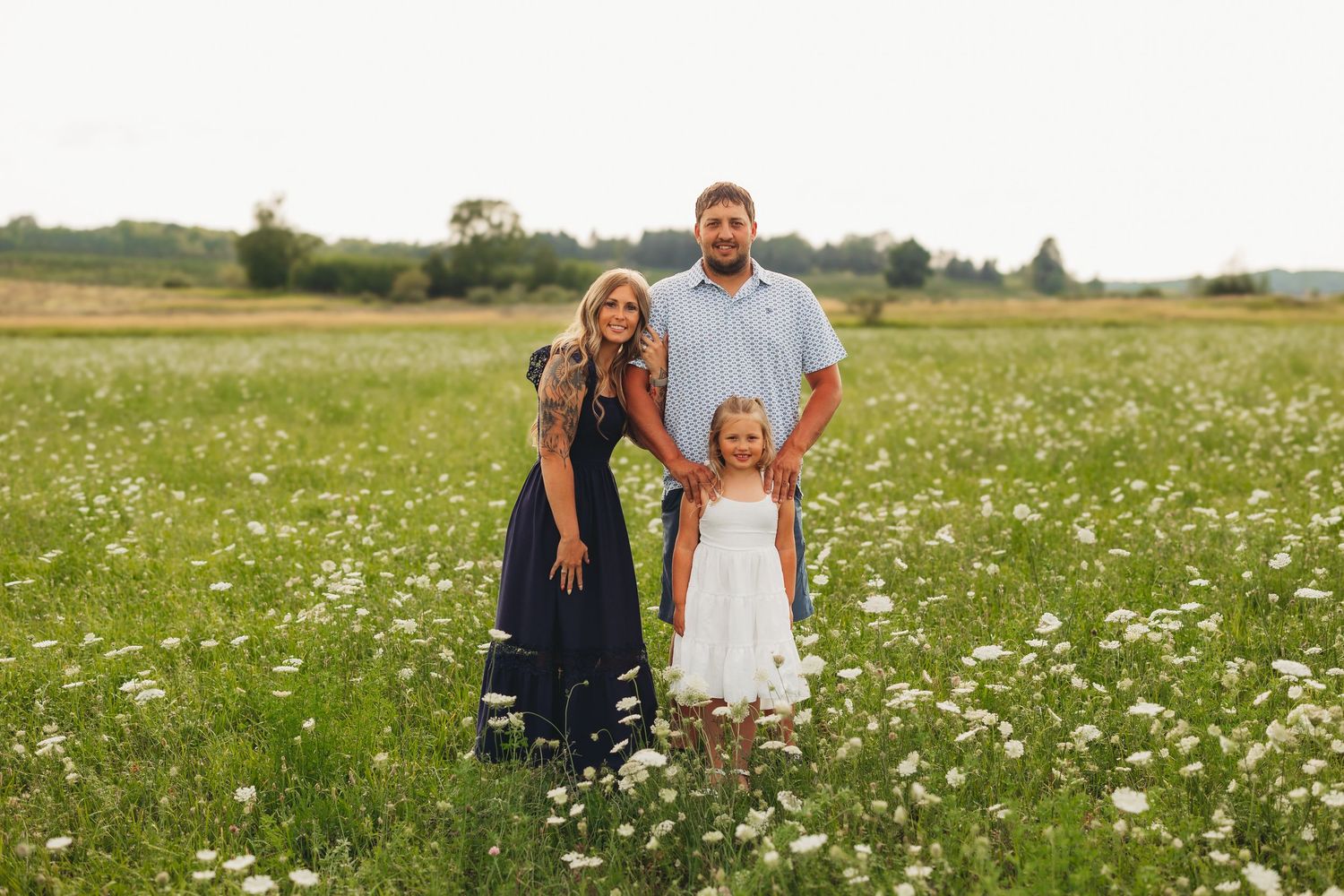 Family of three posing in a wildflower meadow, parents and young daughter in white dress smiling outdoors.