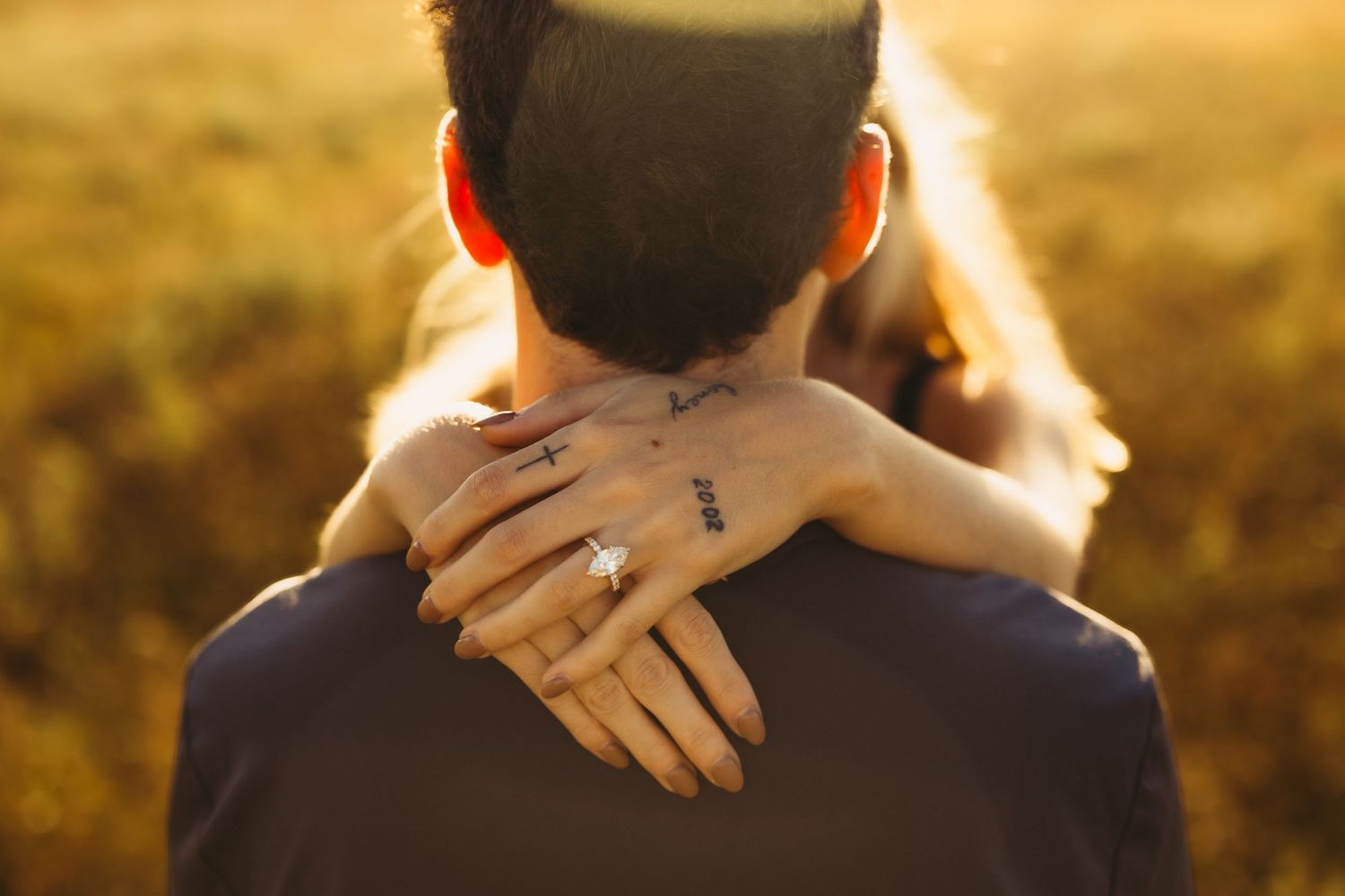 Woman with a sparkling ring and small hand tattoos wraps her arms around a man's neck in a warm embrace against a golden sunset field.