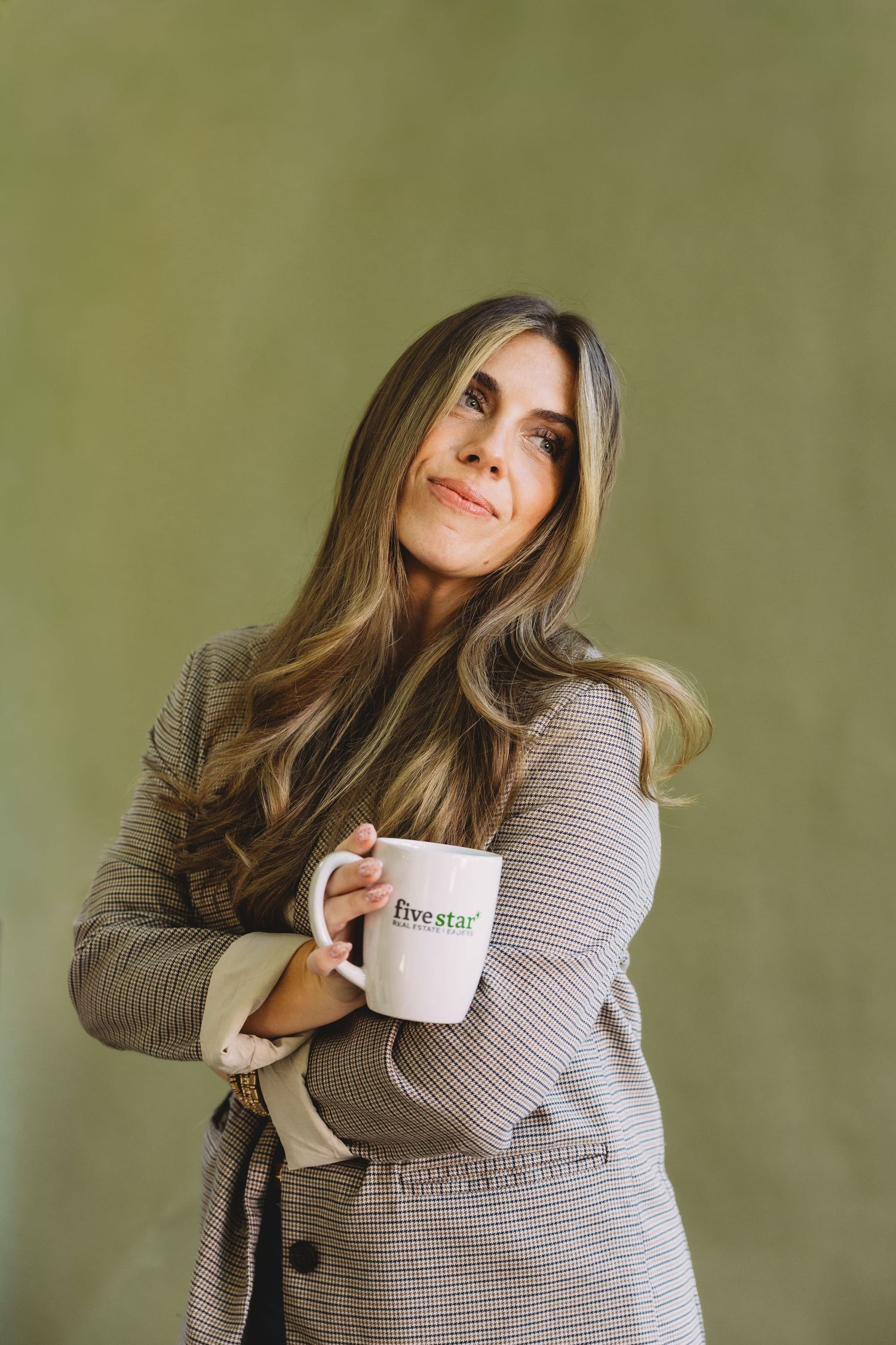 Woman with long wavy hair in a gray tweed blazer smiles while holding a white Five Star coffee mug against a green wall.