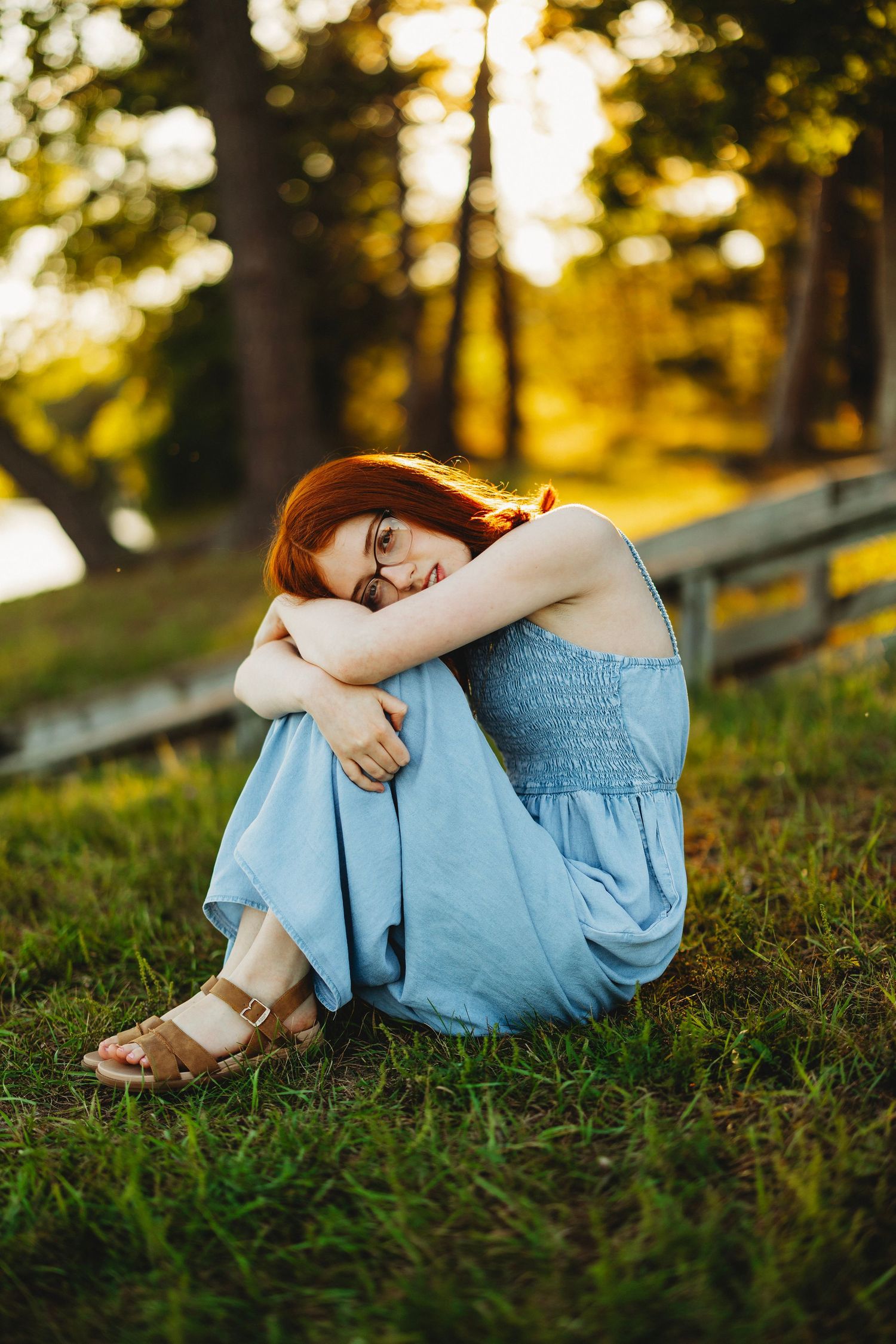 Red-haired woman with glasses in a light blue sundress sits hugging her knees on green grass in warm golden sunset light.