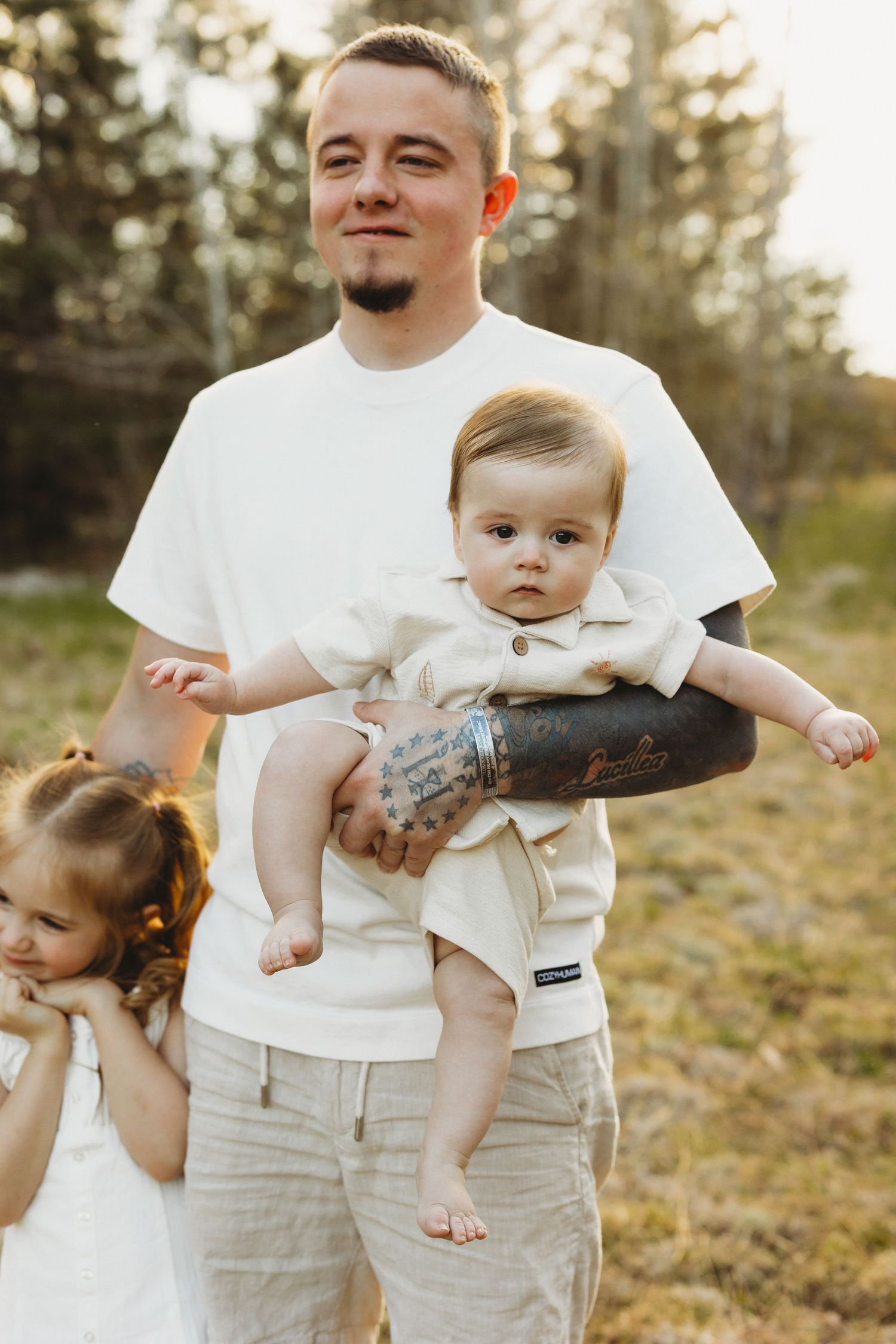 Young tattooed father holding infant son outdoors in sunlit forest setting with toddler daughter beside him.