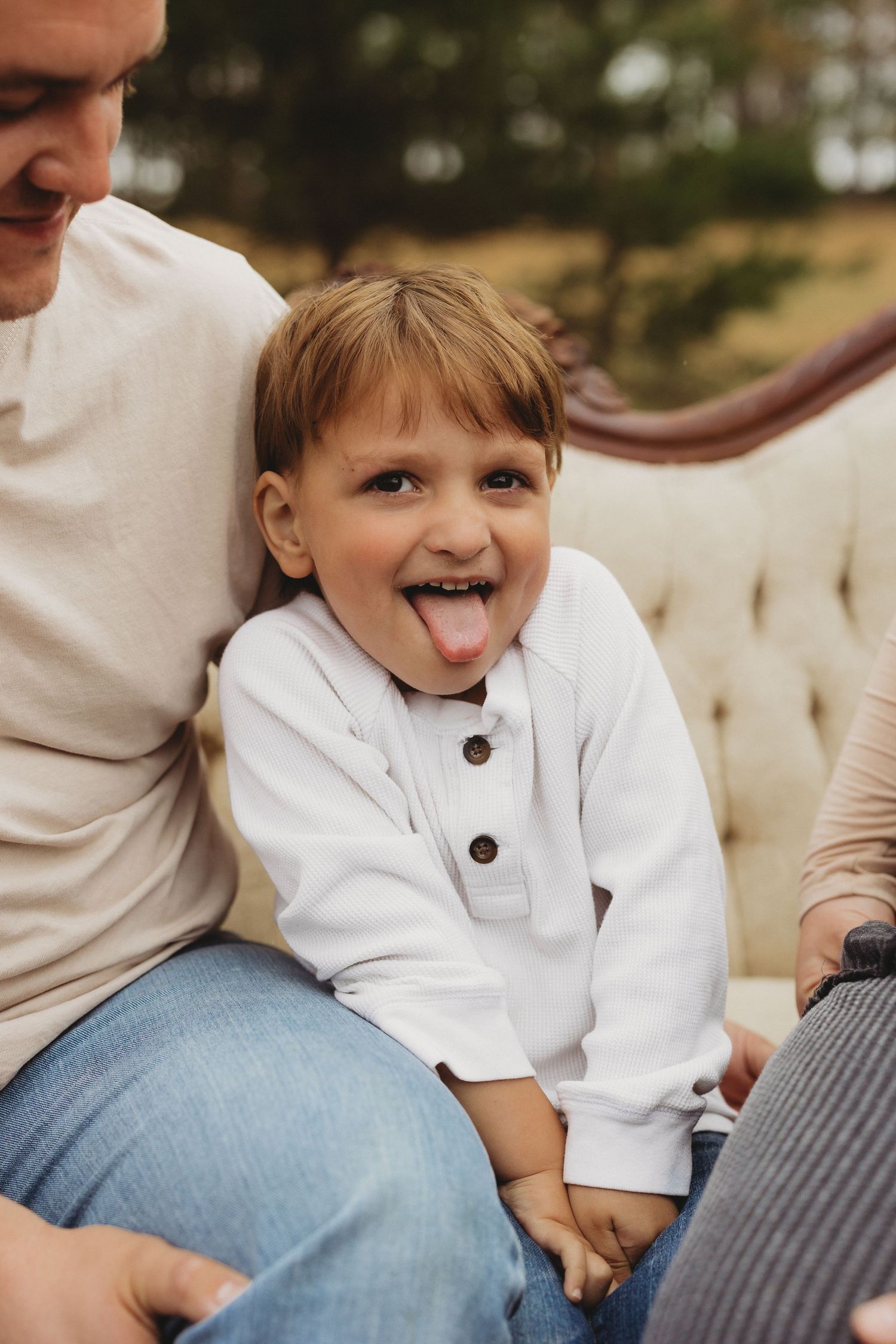 A cheerful young boy sticking out his tongue while sitting on a couch outdoors with an adult.