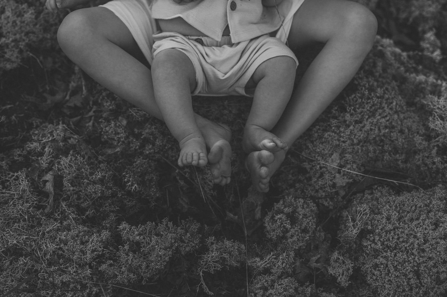 Black and white photo of two children sitting barefoot on mossy ground, legs intertwined in playful togetherness.