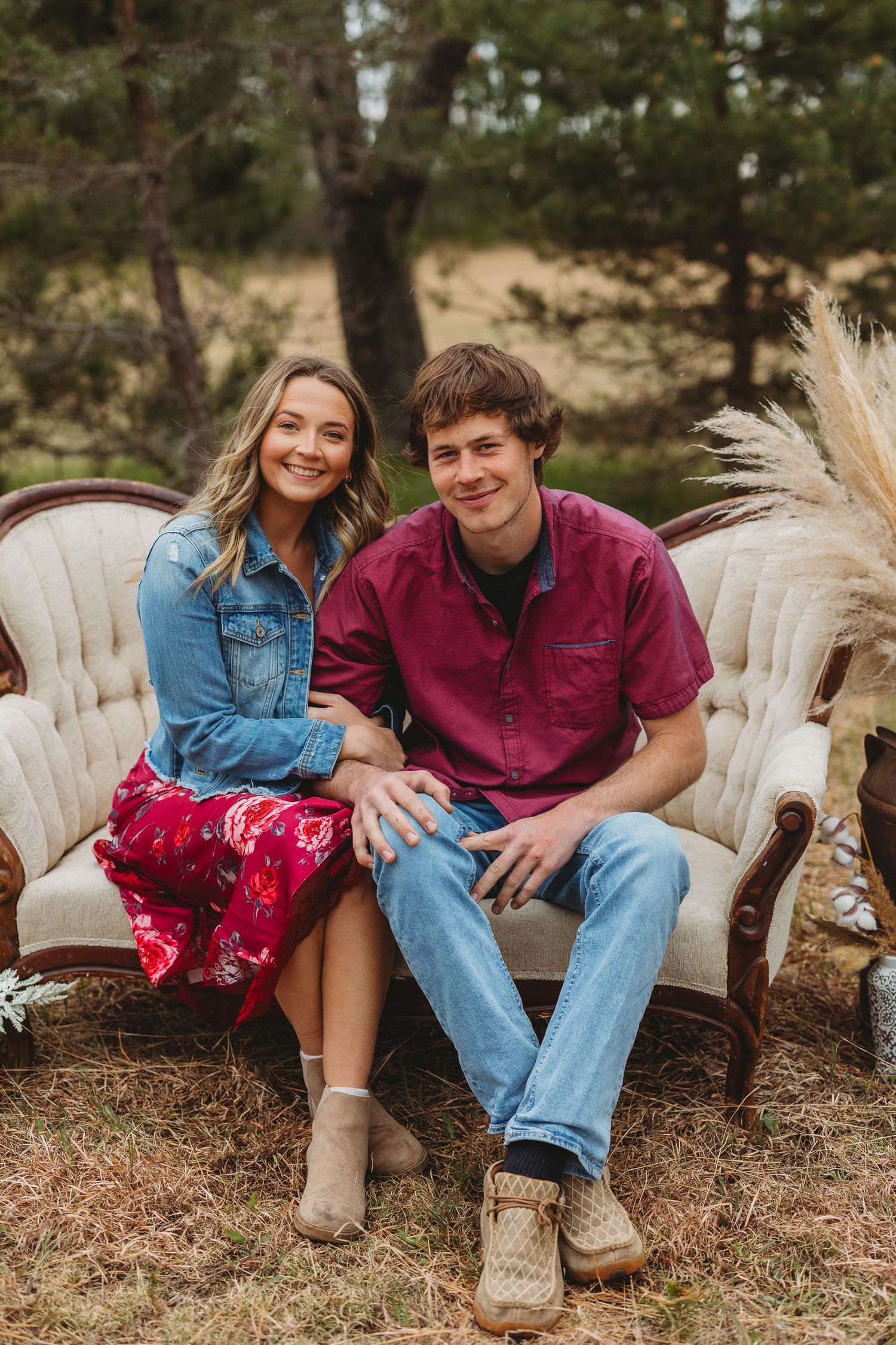 A smiling couple sits together on a vintage sofa outdoors, wearing casual attire in a rustic forest setting.