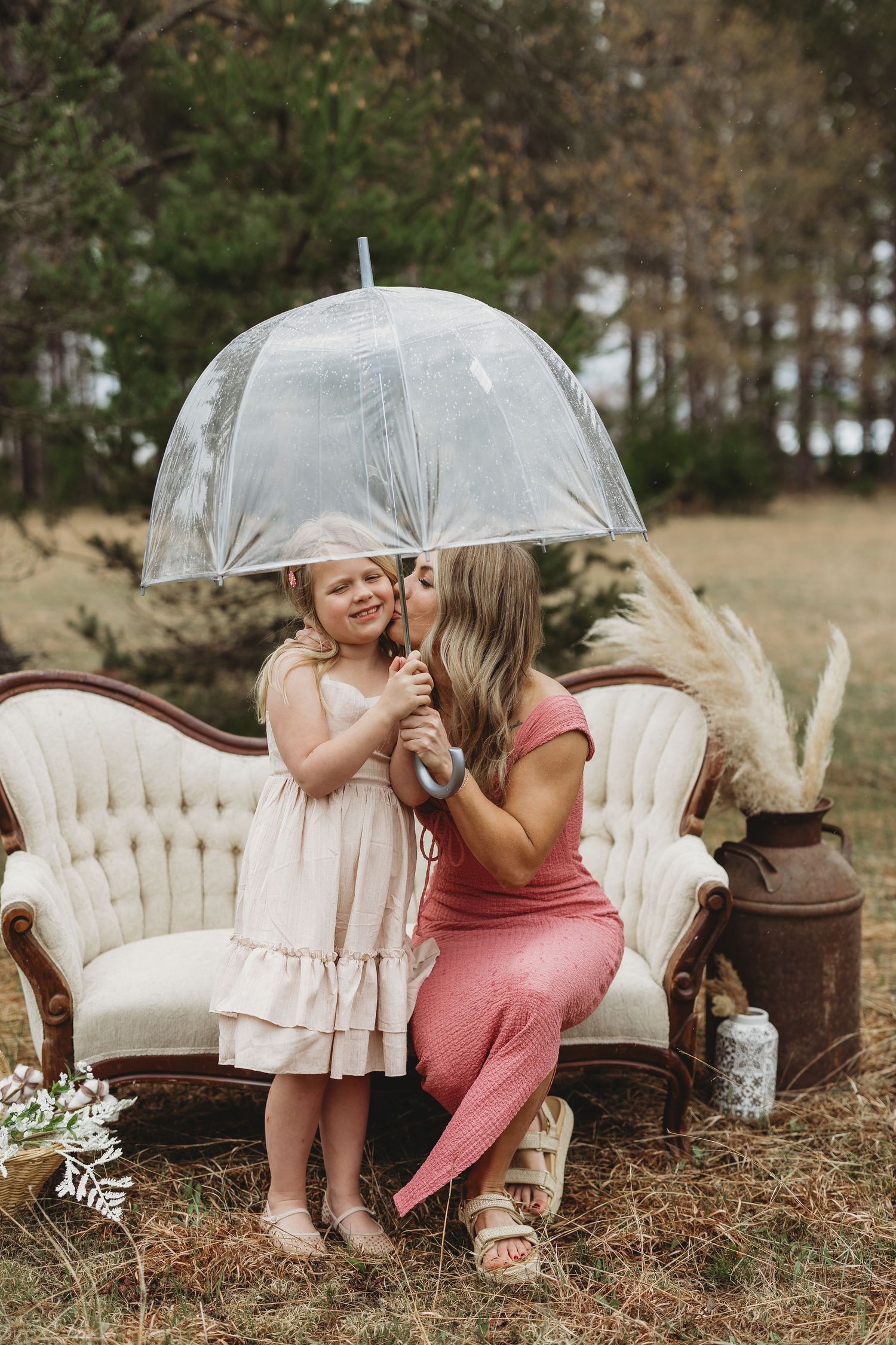 A mother kisses her young daughter holding a clear umbrella on a vintage sofa outdoors.