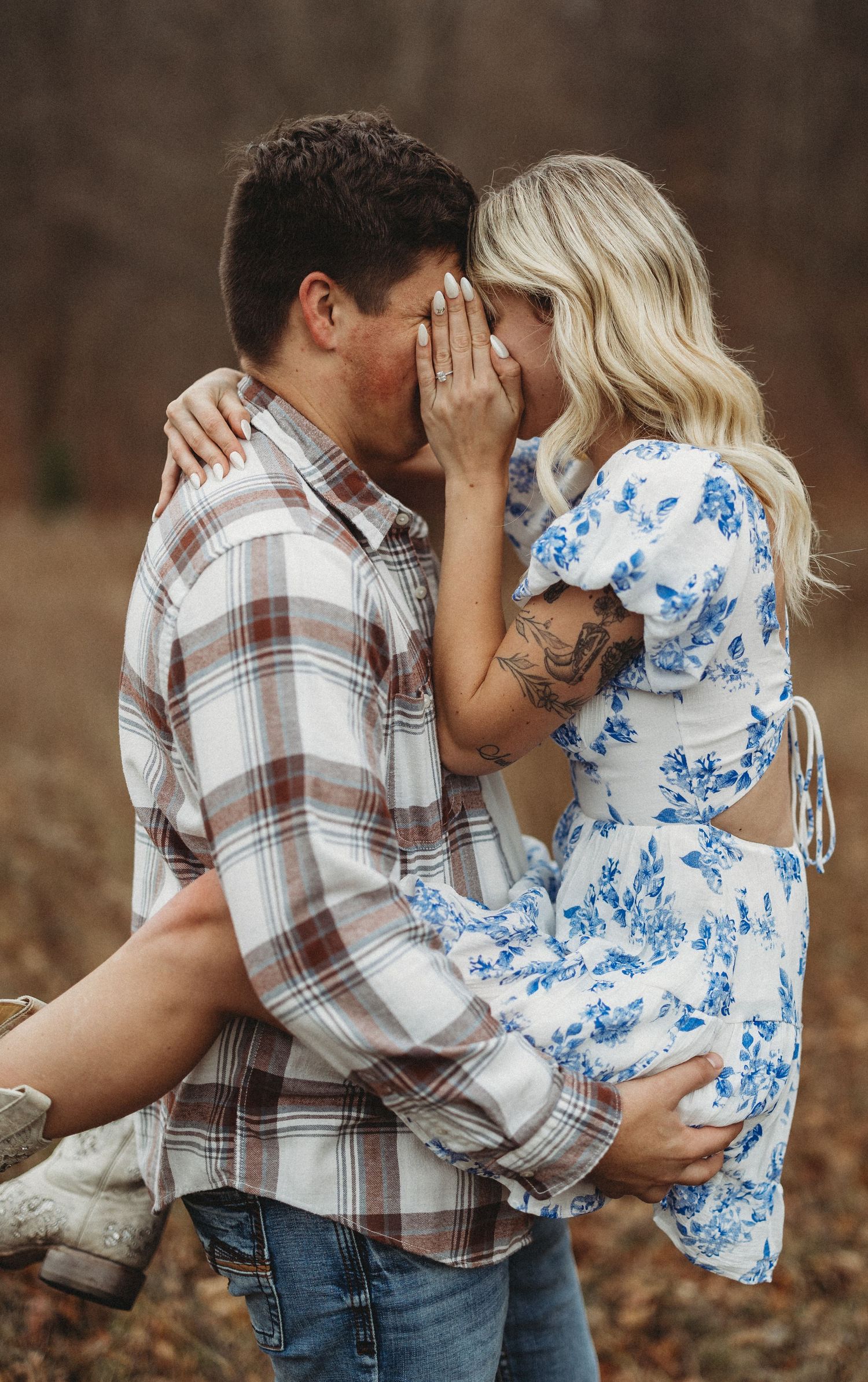 A couple in a plaid shirt and blue floral dress share an intimate embrace while standing among fallen autumn leaves.