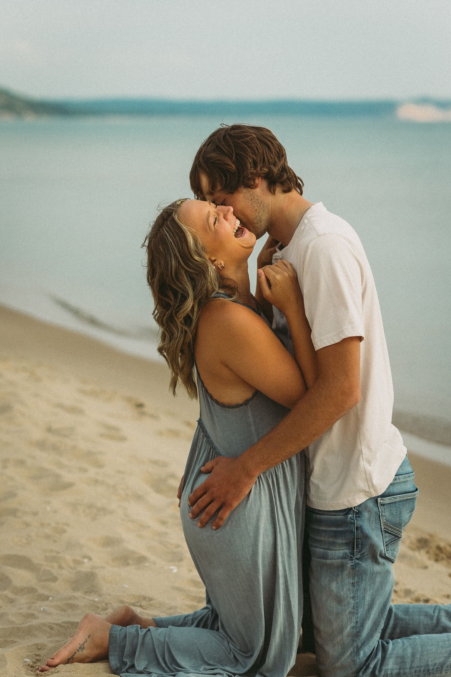 A couple in casual attire sharing tender moments while sitting on a beach at sunset.