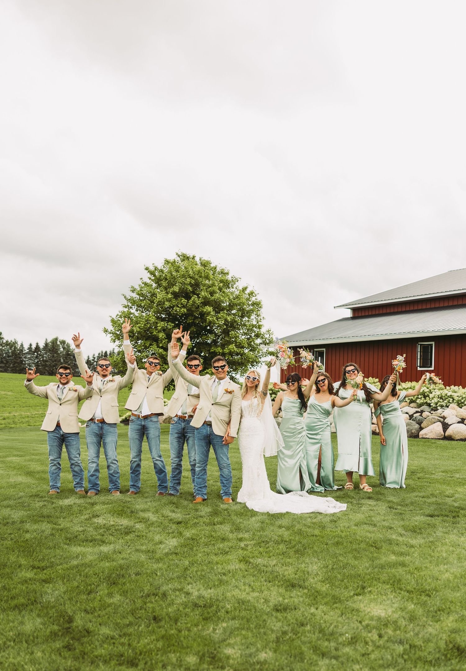 Wedding party celebrates on lawn with arms raised in front of red barn.