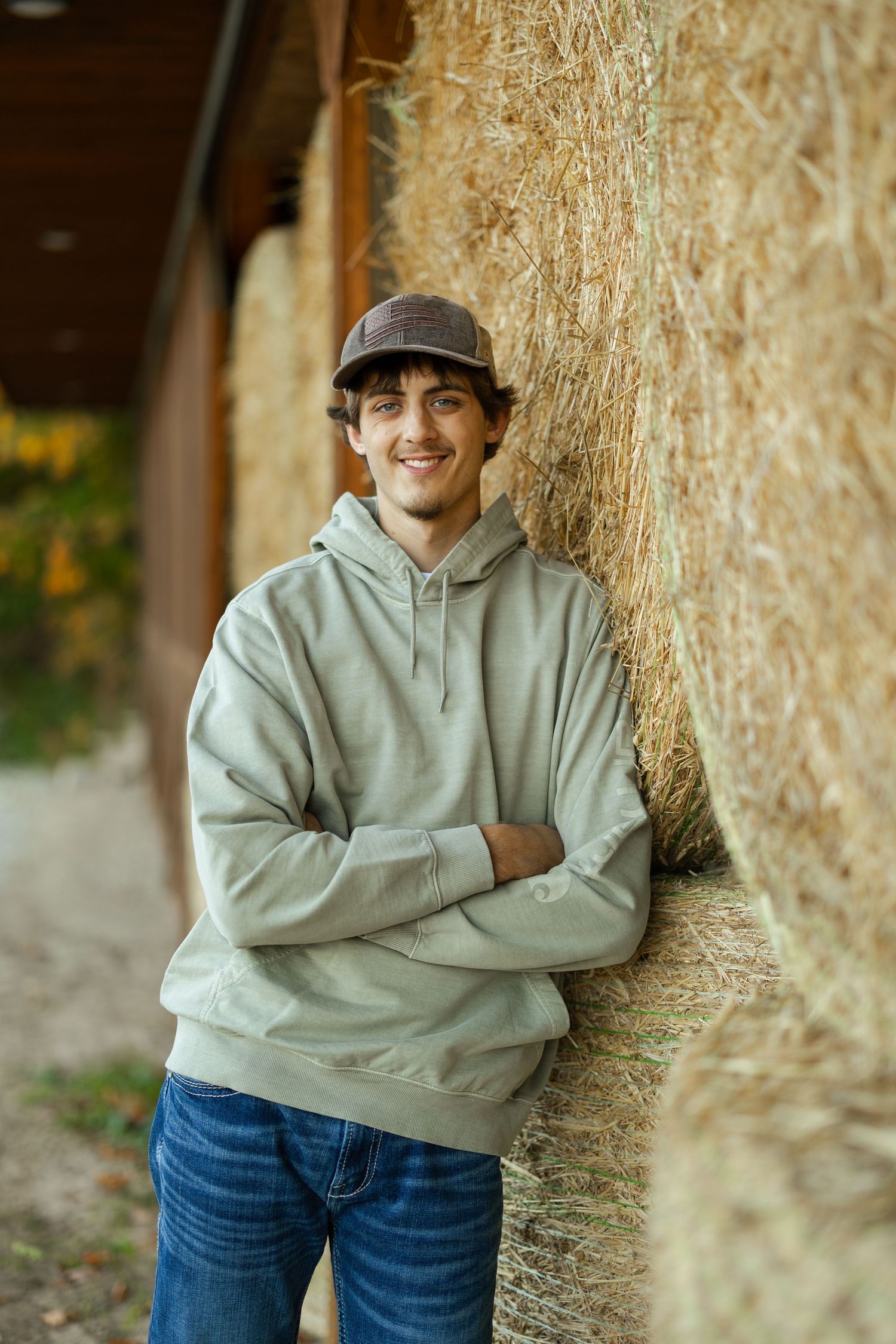 Casual portrait of person in gray hoodie and baseball cap leaning against hay bale wall.