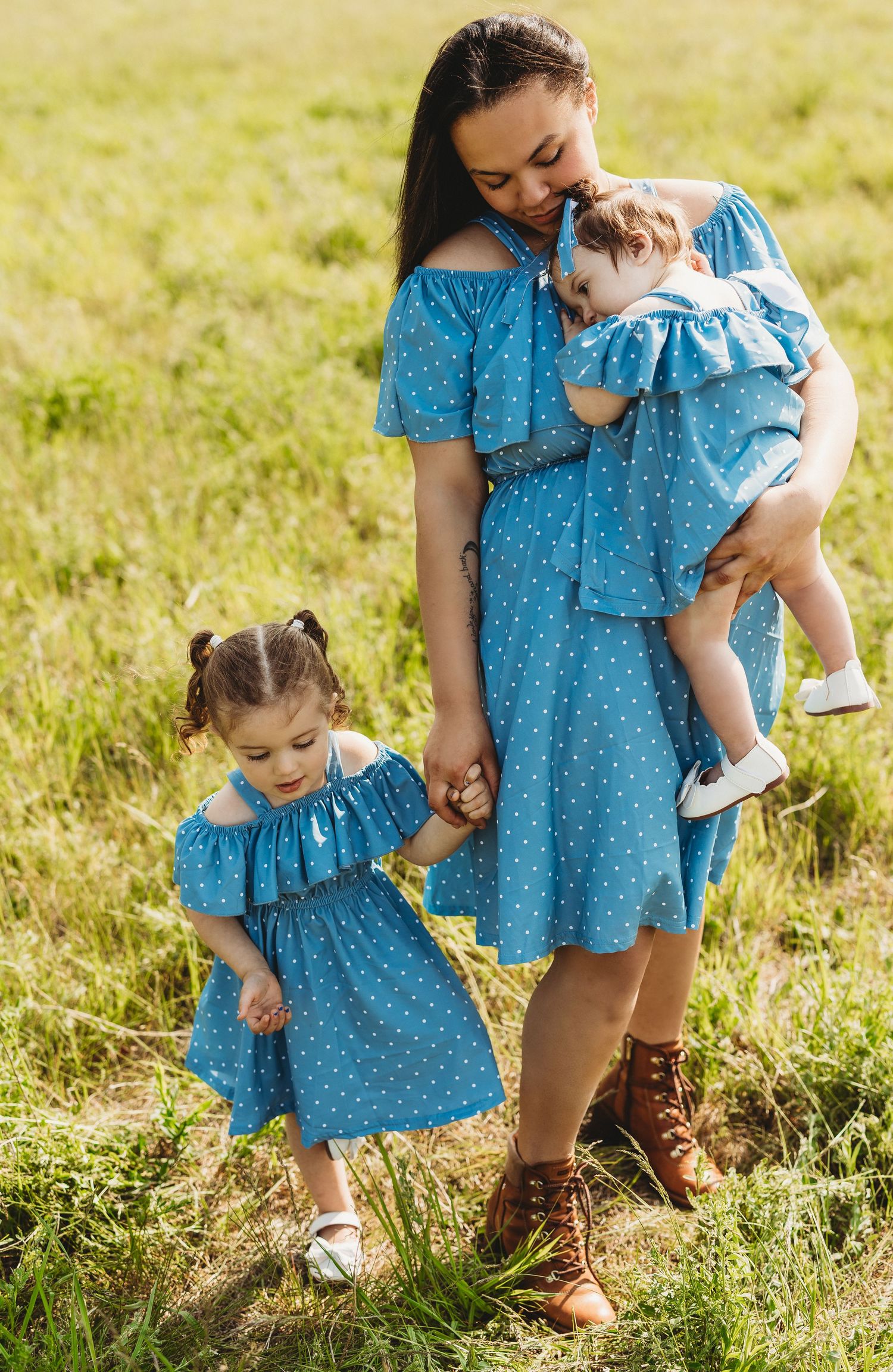 Family wearing matching blue polka dot dresses stands together in sunny meadow with brown leather boots and floral details.