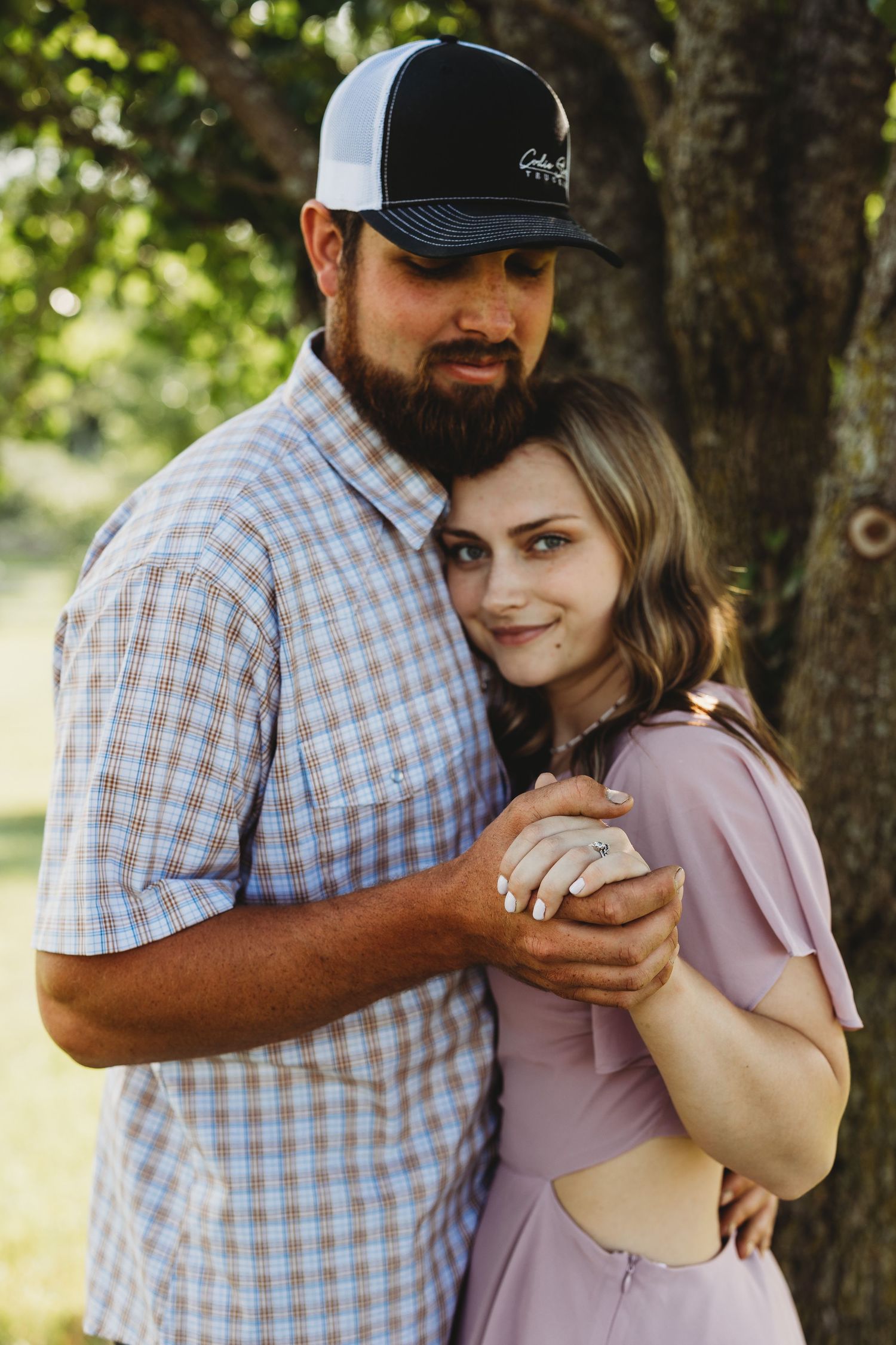 Couple in casual attire embraces tenderly against a tree in a romantic outdoor photoshoot.