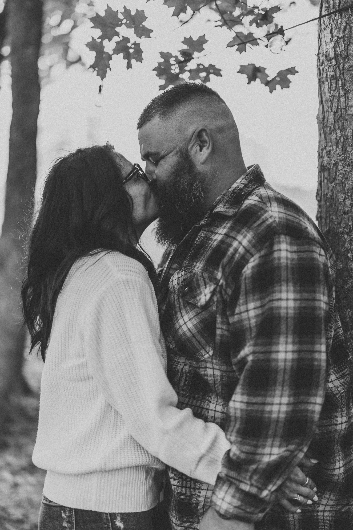 Black and white photo of couple in plaid shirts embracing in a forest setting.