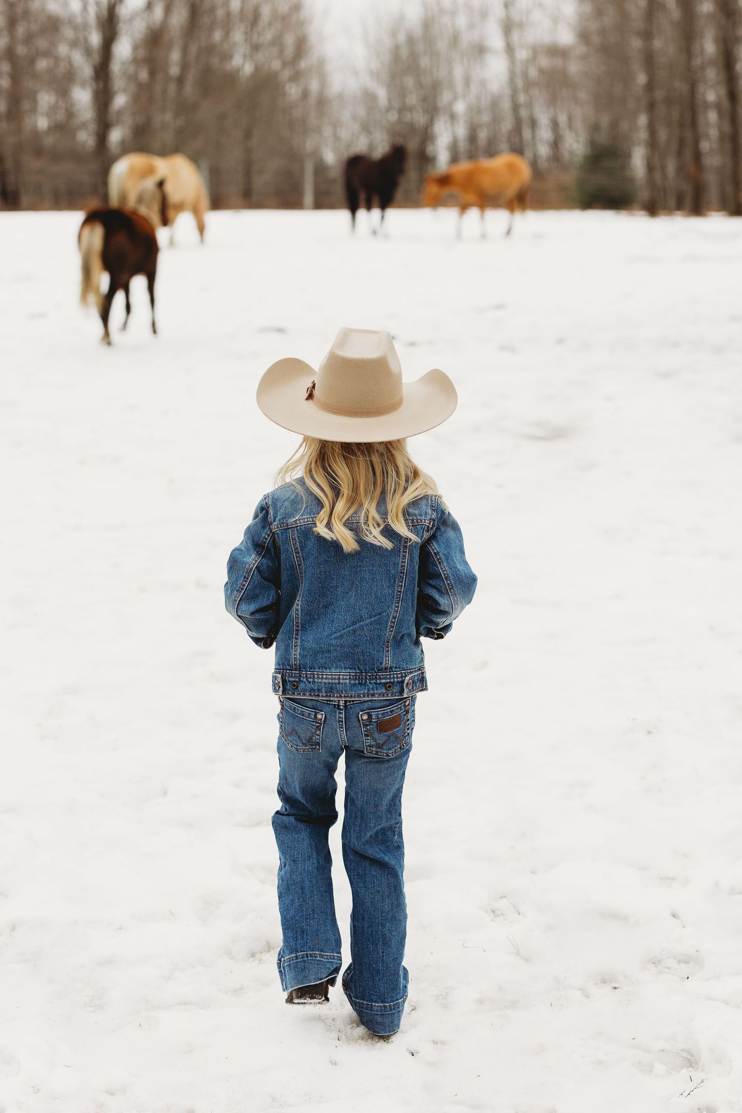 Small child in denim overalls and cowboy hat walking through snowy winter landscape with horses visible in background.