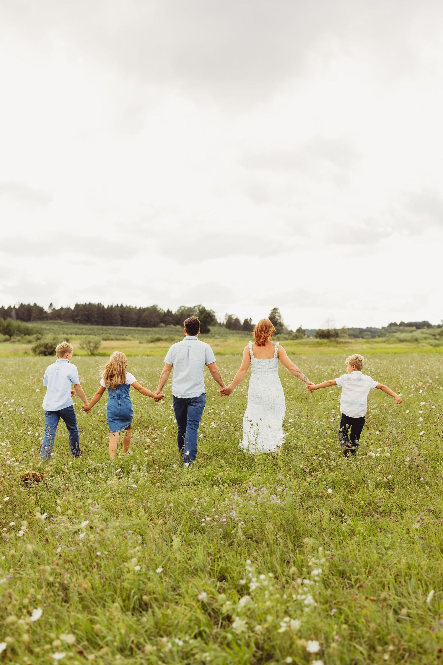 Family of five holding hands walking away through a green wildflower meadow under a cloudy sky, with trees along the distant horizon.