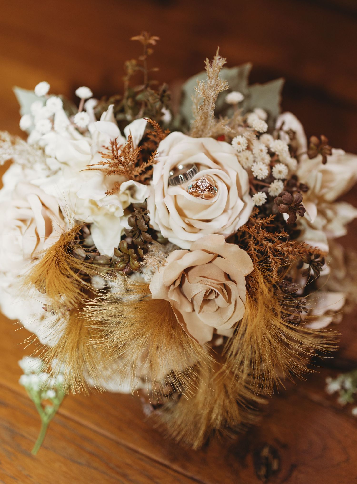 Rustic bridal bouquet of cream roses, dried wildflowers and golden feathers with wedding rings nestled in the blooms on a wooden table.