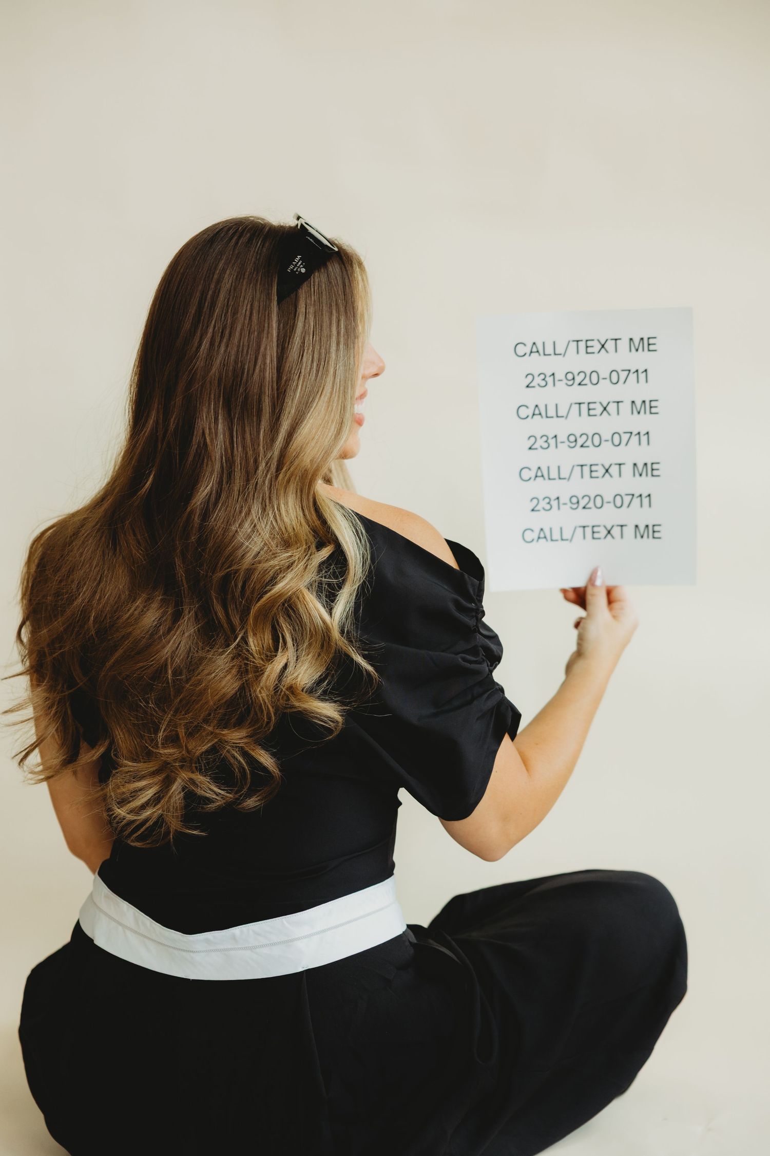 Woman with long wavy hair seated facing away, holding a sign repeating Call/Text Me with a phone number.