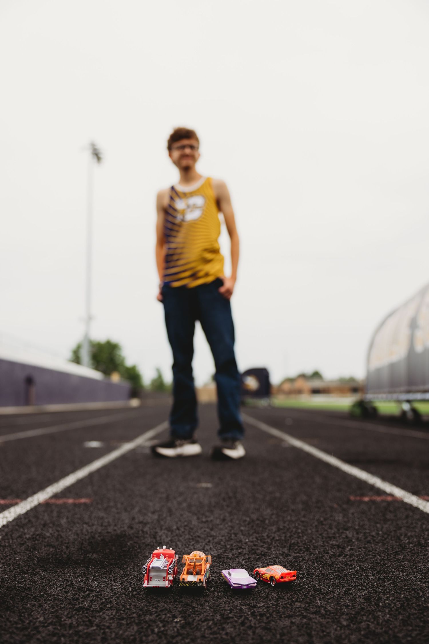 Smiling runner in a yellow tank top and jeans stands on a track behind four small toy cars lined up in the foreground.