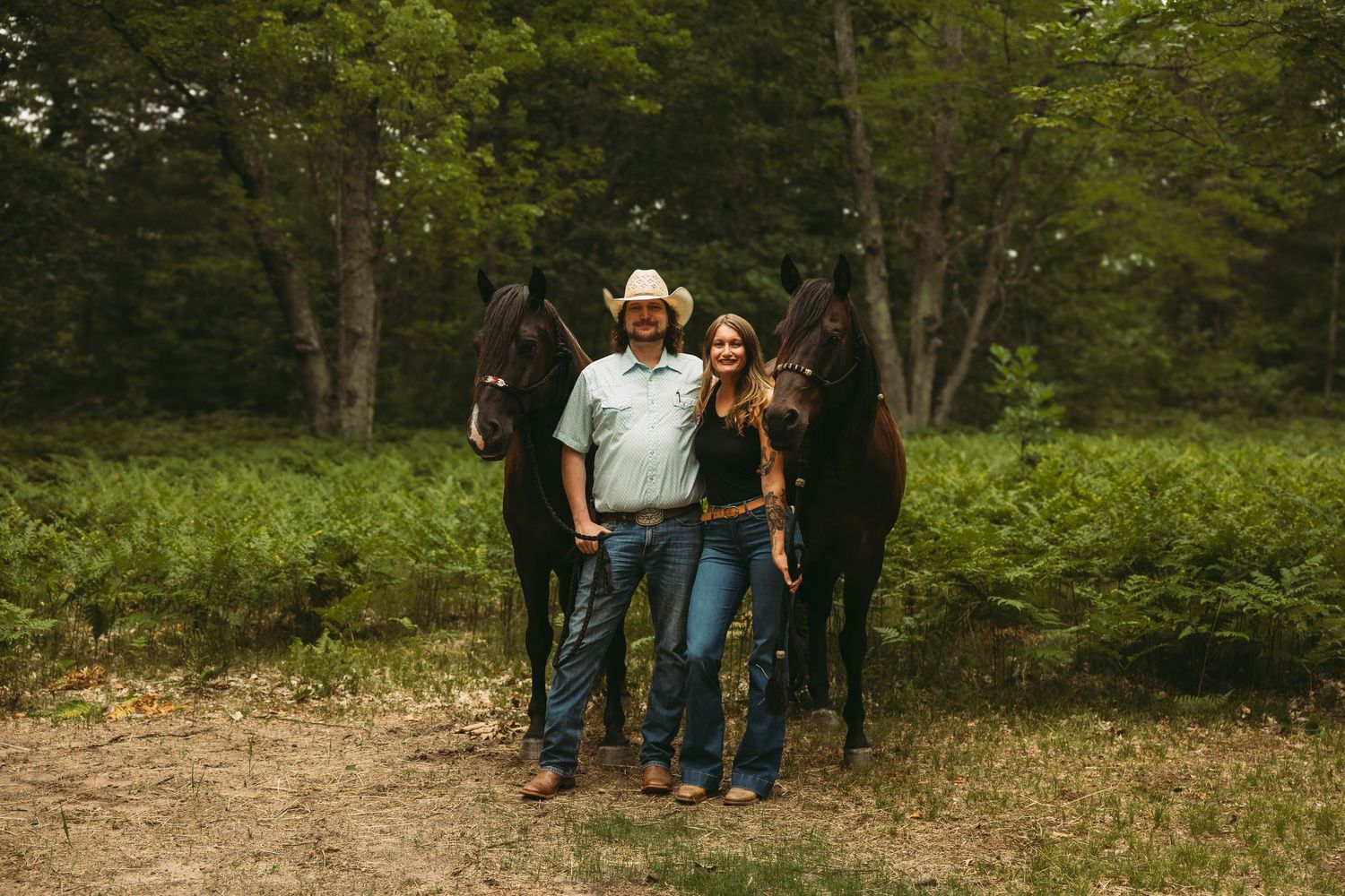 Couple in cowboy hats posing with two black horses in a lush green forest clearing.