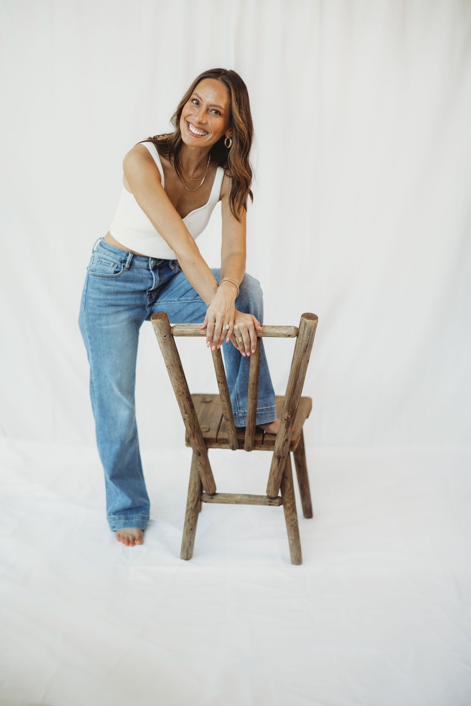 Smiling woman in white tank top and blue jeans leaning on a rustic wooden chair against a white backdrop.