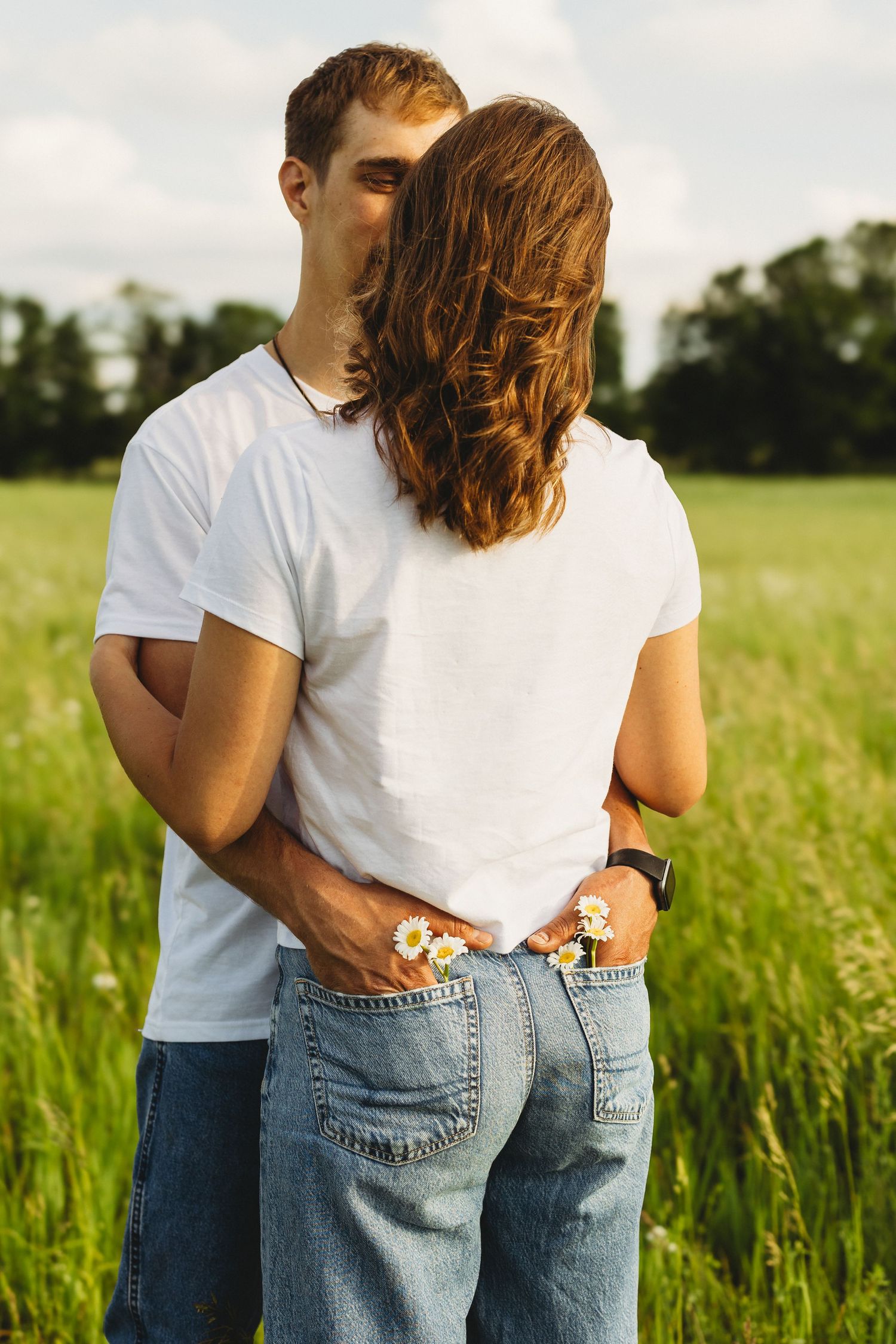 Couple embracing in a green field, wearing white t-shirts and jeans, holding daisies behind their back.