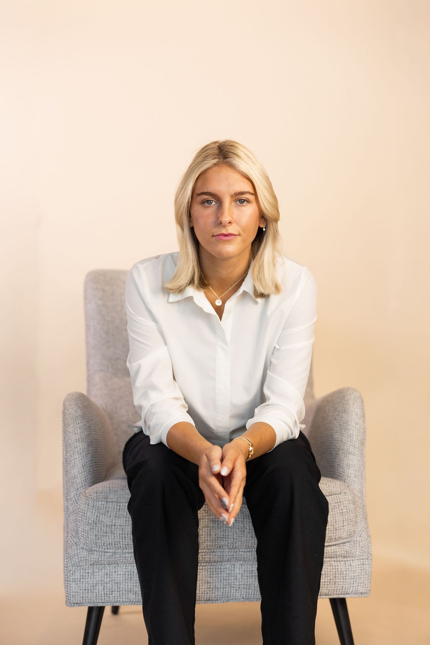 Blonde woman in white shirt and black pants sitting in gray armchair against beige background.