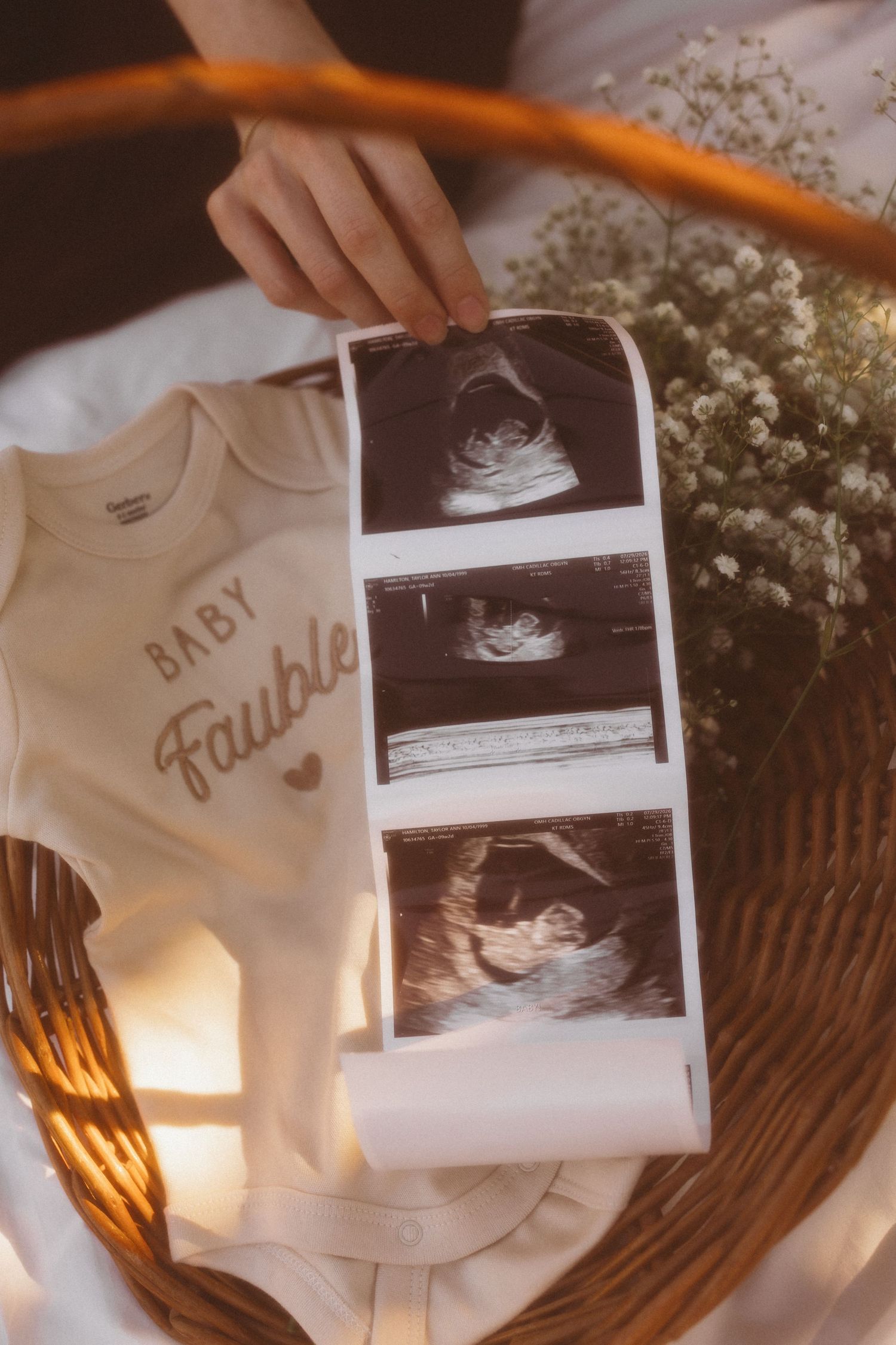 Hand holding ultrasound photos beside a white Baby Faubl onesie and dried flowers in a wicker basket.