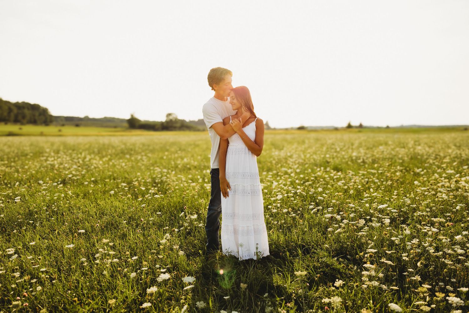Couple embracing in a sunlit wildflower field, woman in white dress, golden hour romantic outdoor photography.
