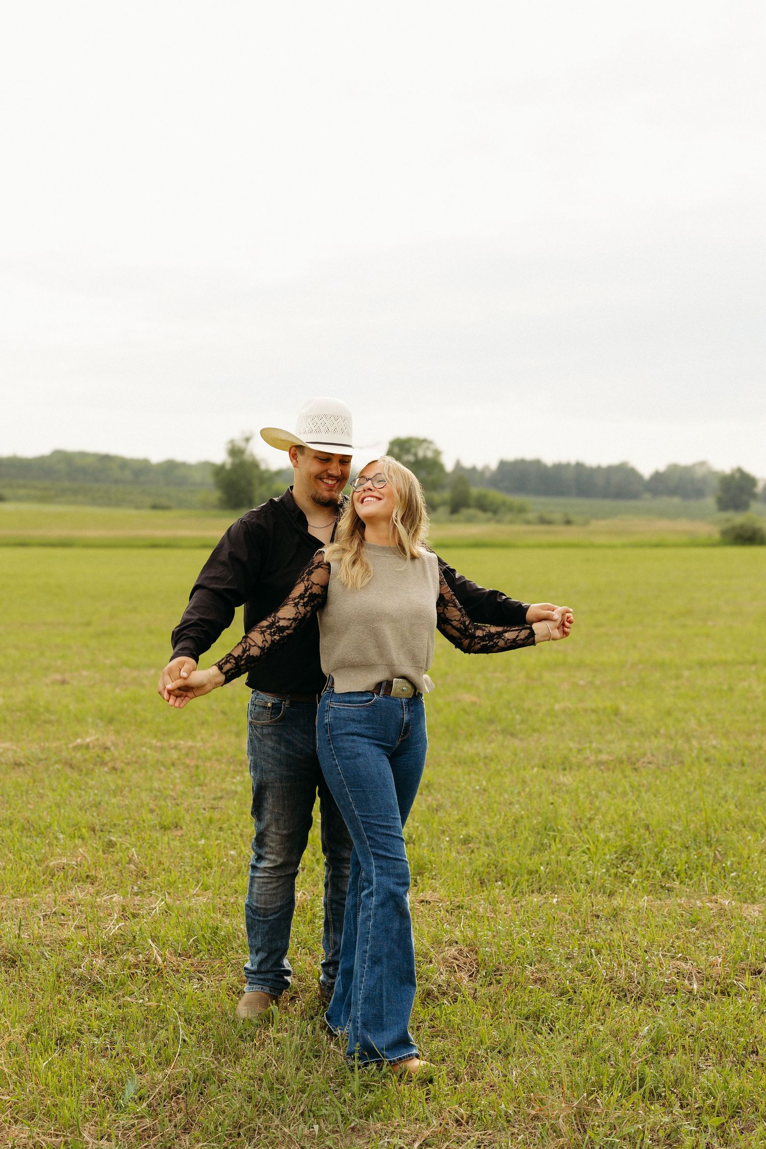 A happy couple dances in a green field, woman in flare jeans and white hat, man in dark jacket and jeans.