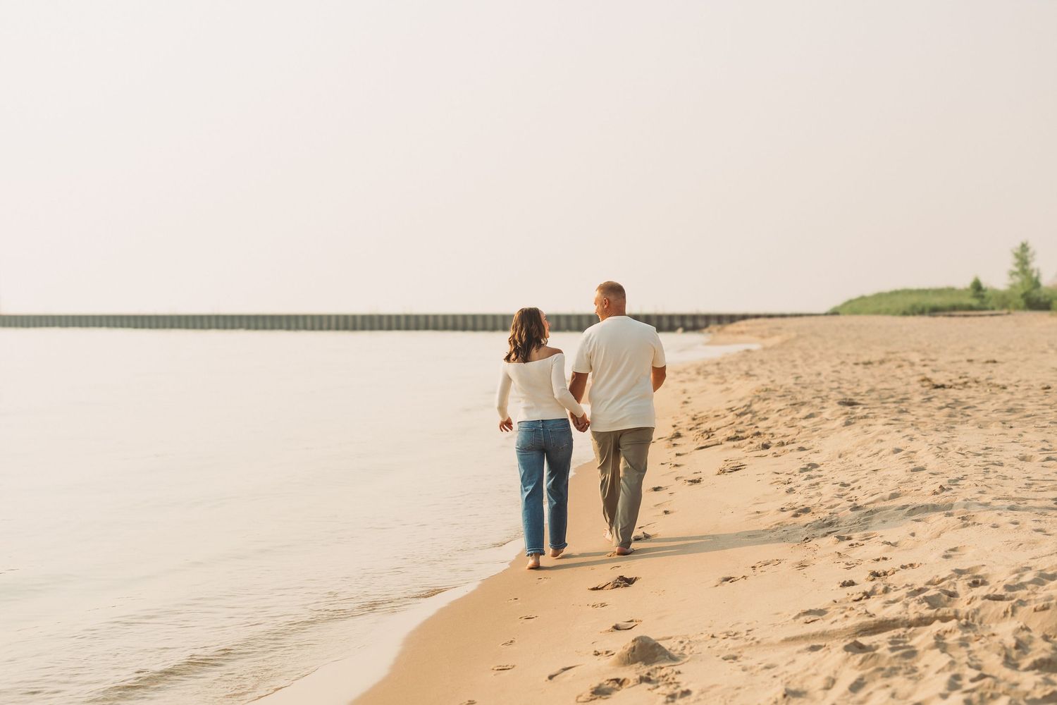Couple walking hand in hand along a sandy beach shoreline with a pier in the background on a hazy day.