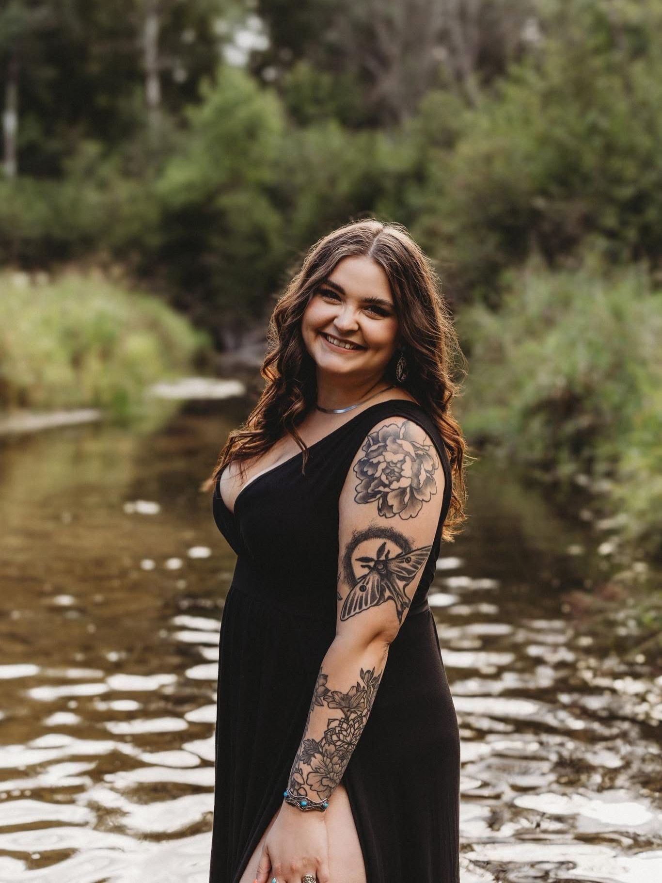 Smiling woman with tattoos in black dress standing in a shallow creek surrounded by lush greenery.