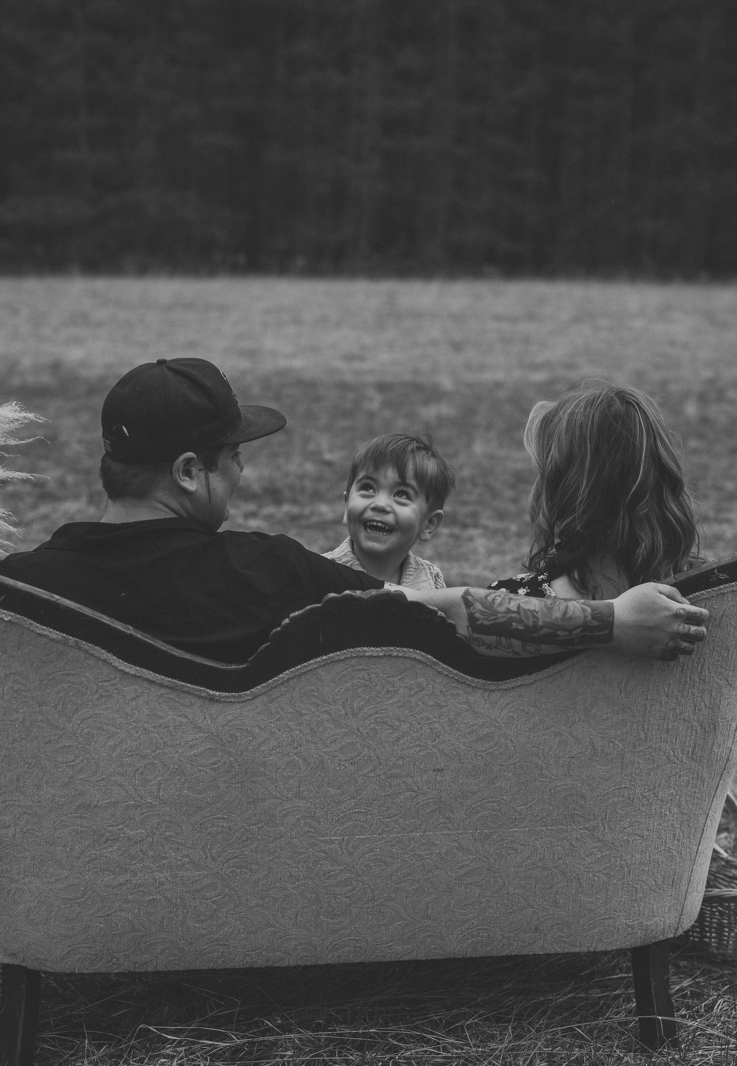 Black and white photo of a man and two children sitting together on a vintage sofa outdoors, sharing a joyful moment.