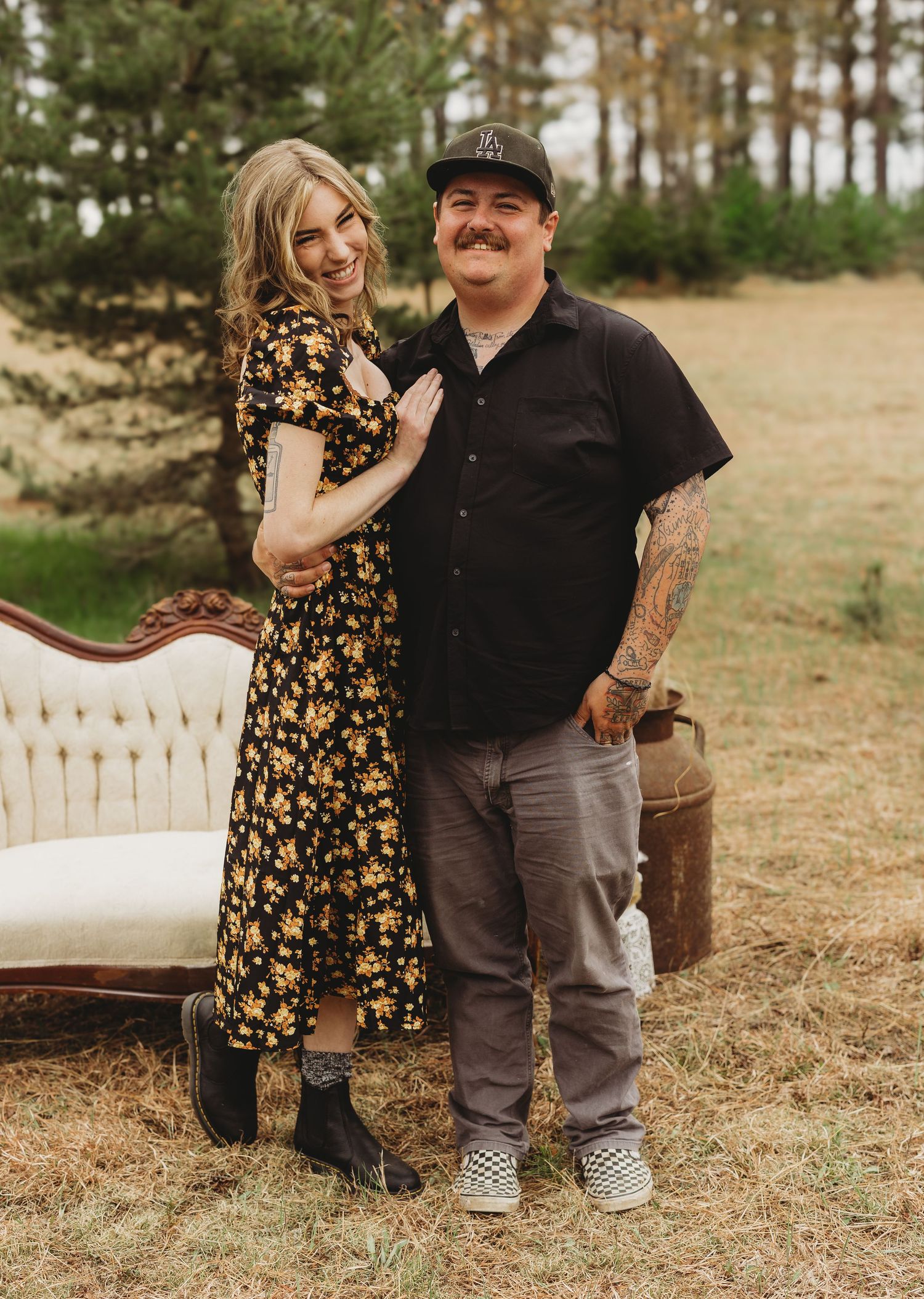 Couple posing outdoors near vintage sofa in field, woman in floral dress, man in black shirt with tattoos.