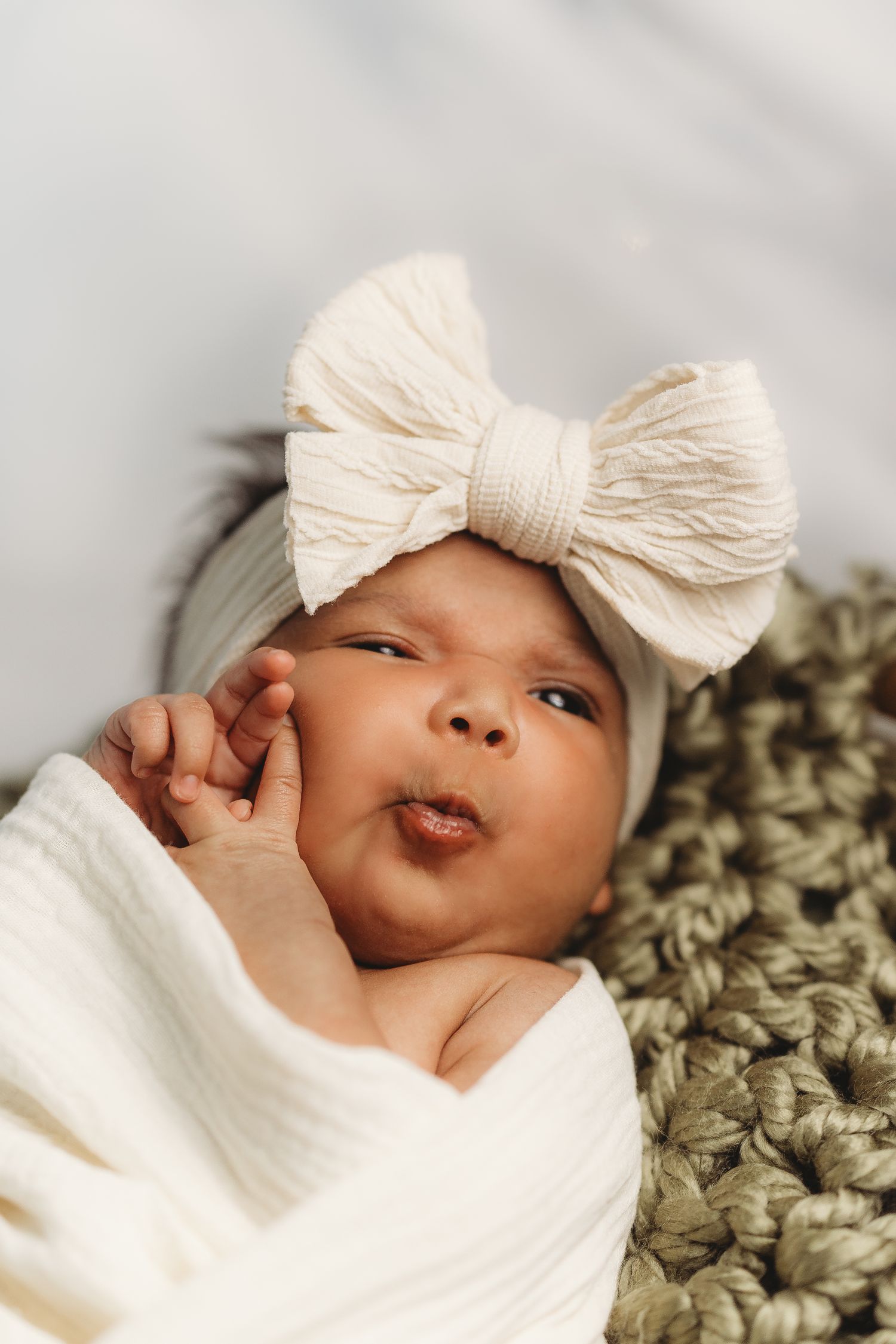 Newborn baby wrapped in white swaddle wearing cream bow headband lying on knit blanket.