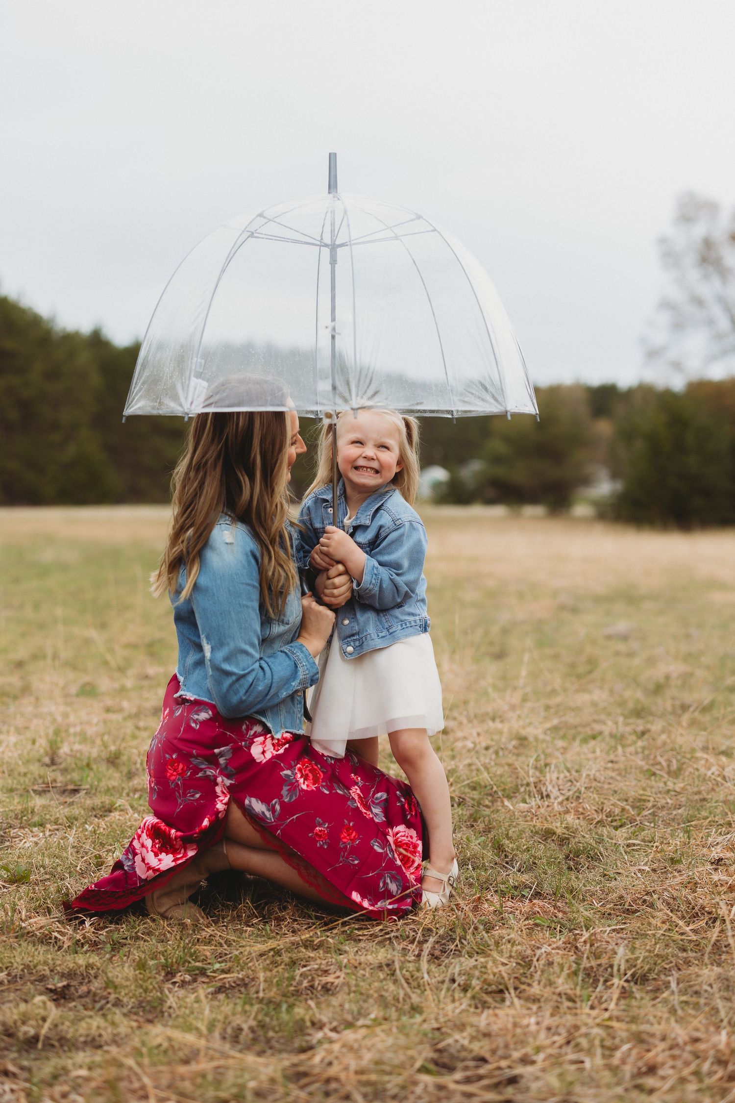 A mother kneels with her smiling toddler under a clear bubble umbrella in a field, both wearing denim jackets.