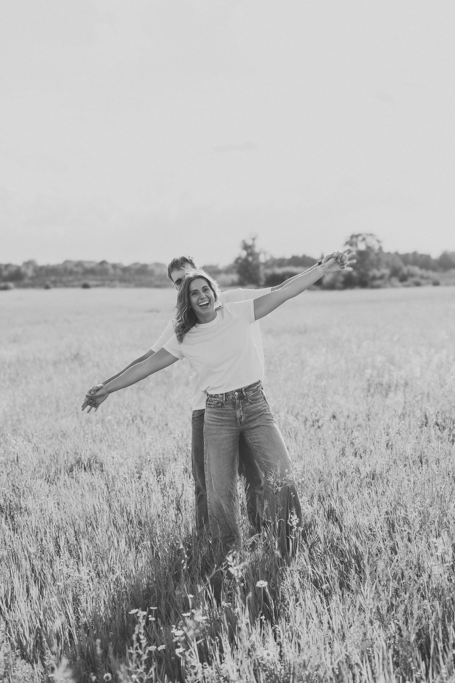 A joyful woman stands in a field with arms outstretched, wearing jeans and a white tee, in black and white.