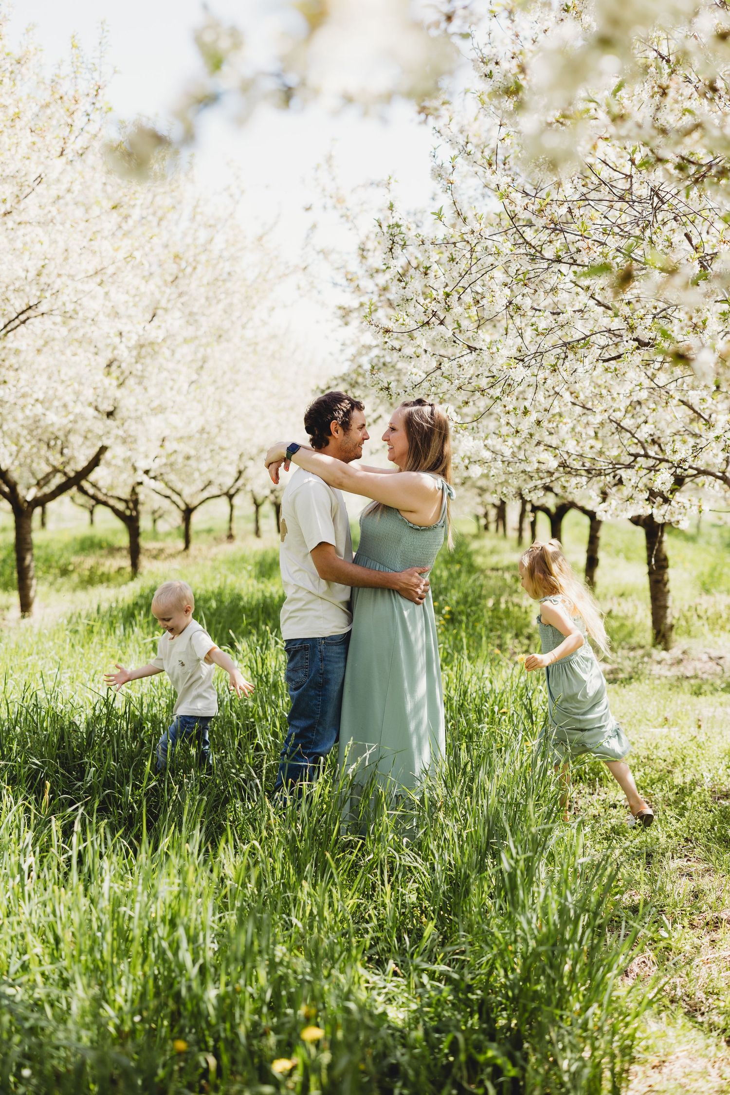 A couple embraces and kisses in a blooming orchard while two young children play around them in tall green grass.