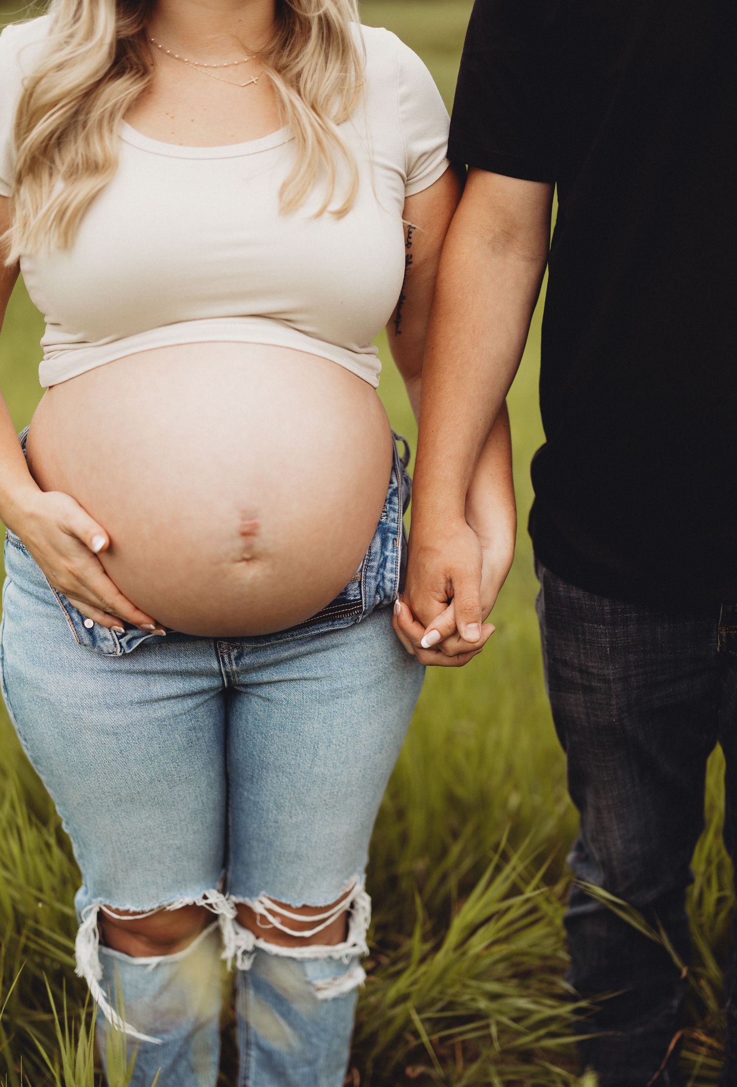 Close-up of pregnant woman's bare baby bump holding partner's hand in field.