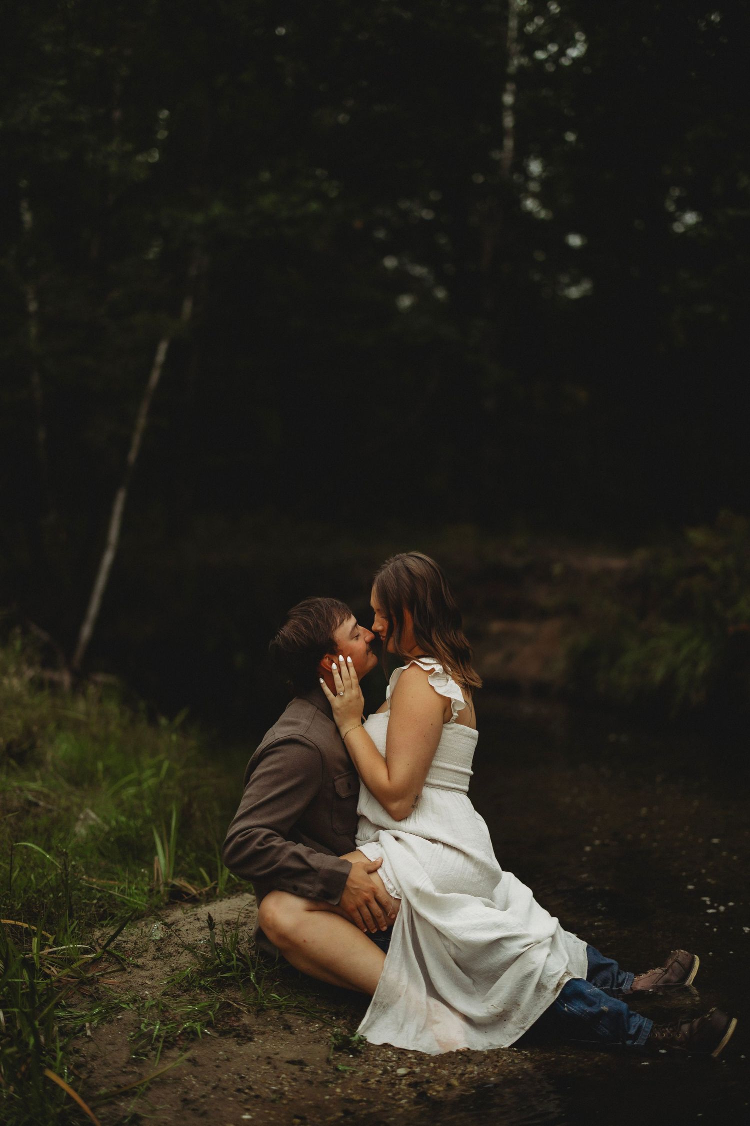 Couple in a white dress and brown shirt share an intimate kiss while sitting on a mossy forest bank beside a quiet stream.