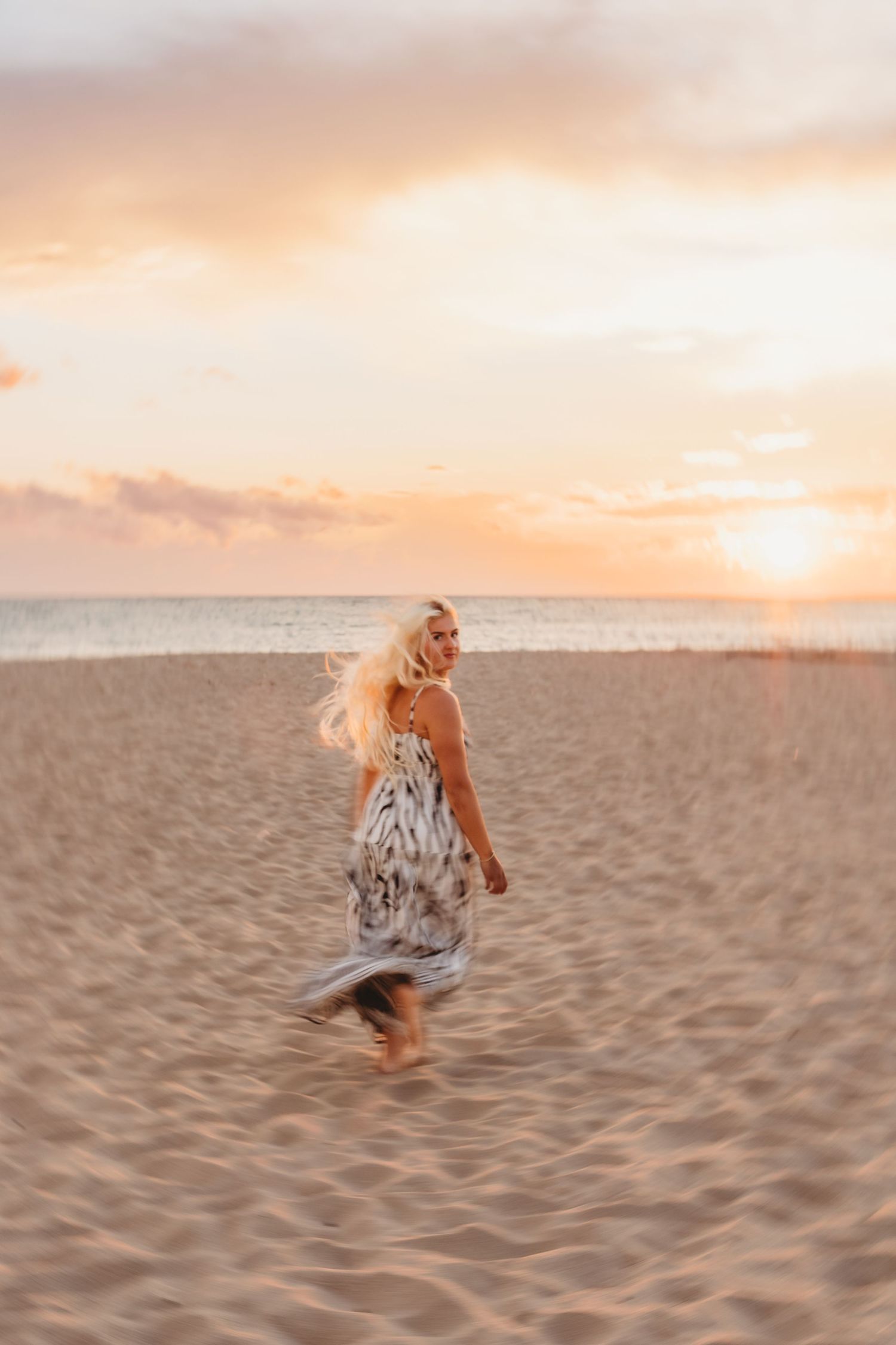 Woman in a flowing patterned dress twirls on a sandy beach at sunset, with golden light, soft clouds and calm ocean waves behind her.