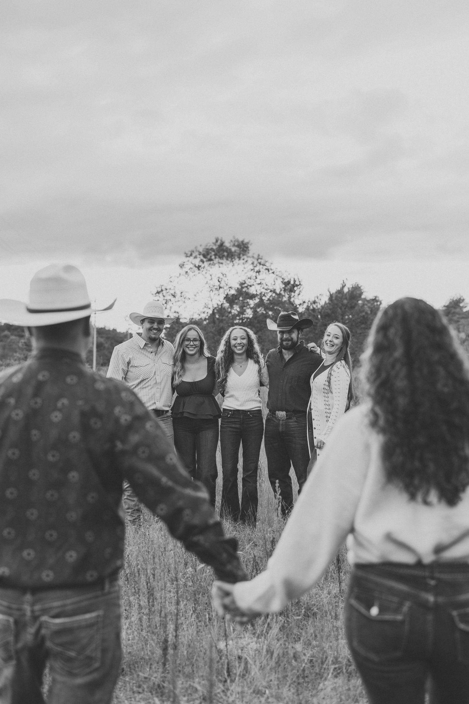 Black and white photo of a family in cowboy hats and casual clothes holding hands in a circle in a grassy field under a cloudy sky.