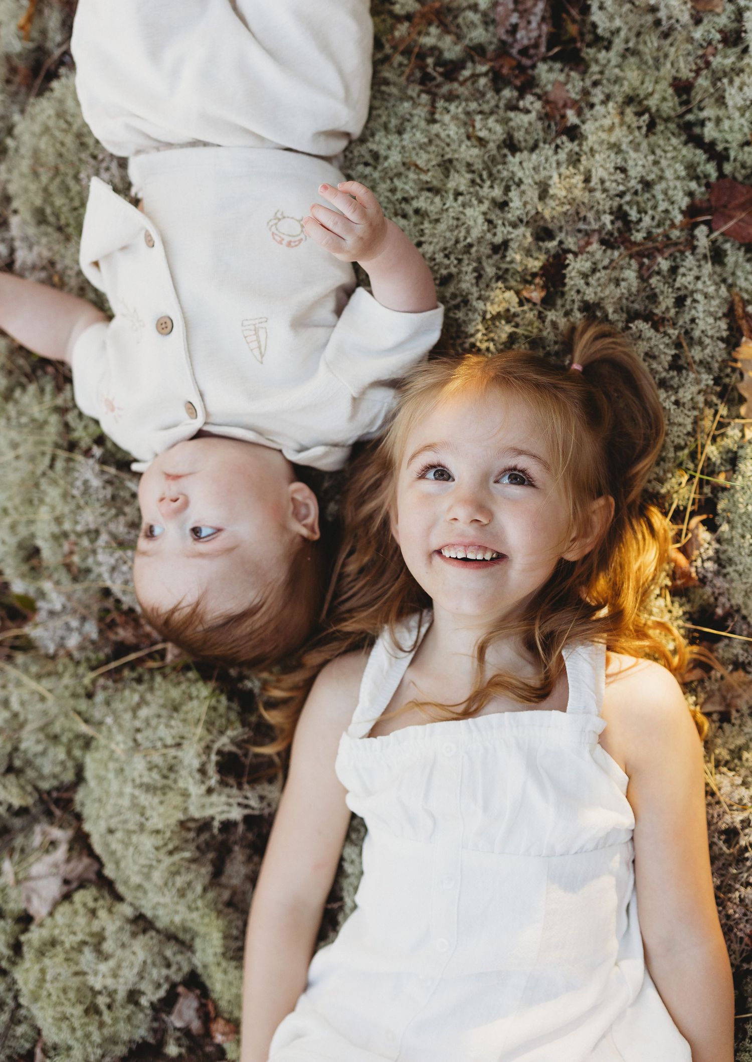 Two young siblings lying on moss, smiling girl in white dress and baby in cream outfit looking up.
