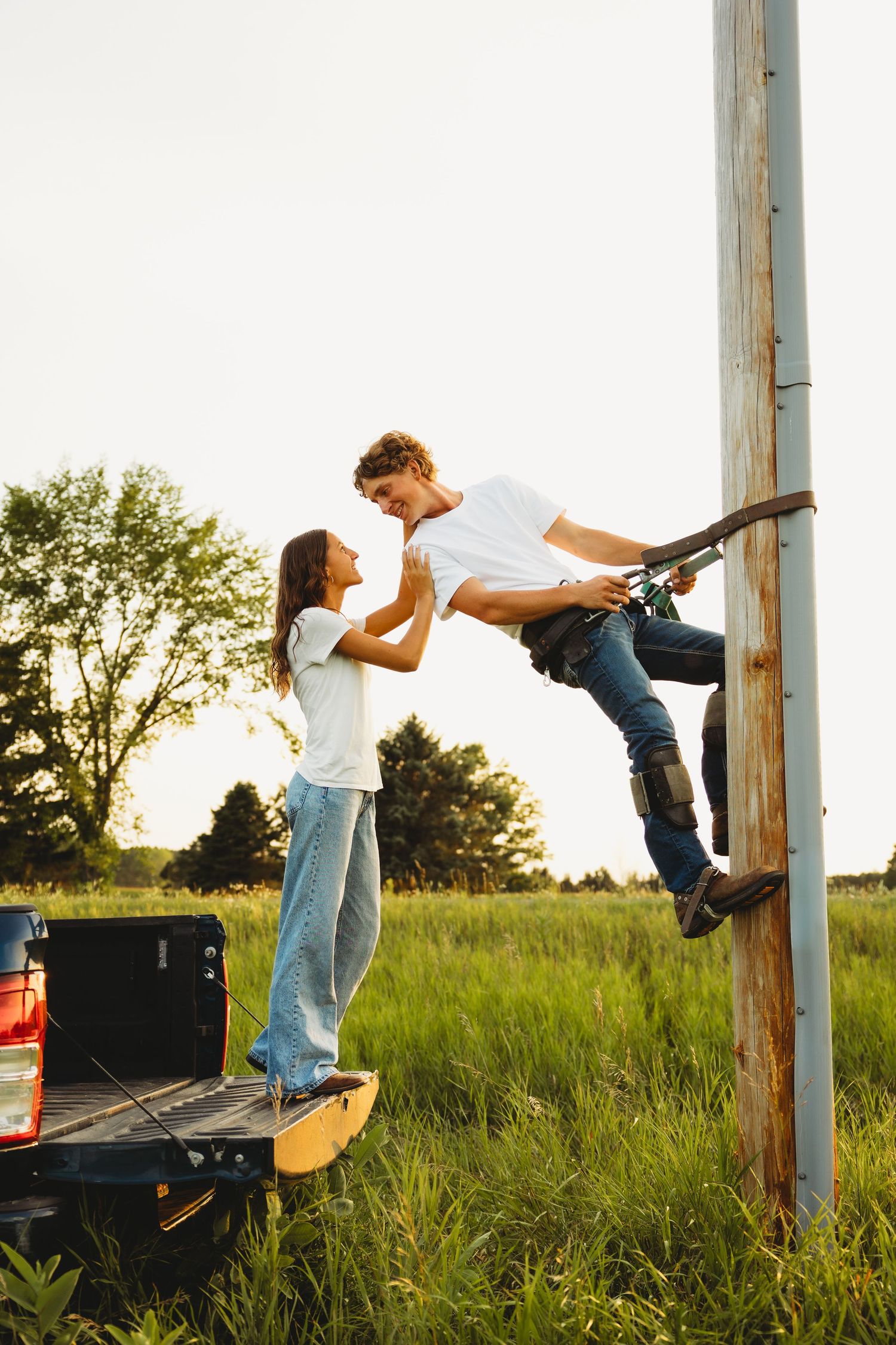 A young couple shares a romantic moment as the man climbs a wooden pole while the woman stands on a truck bed in a green field.