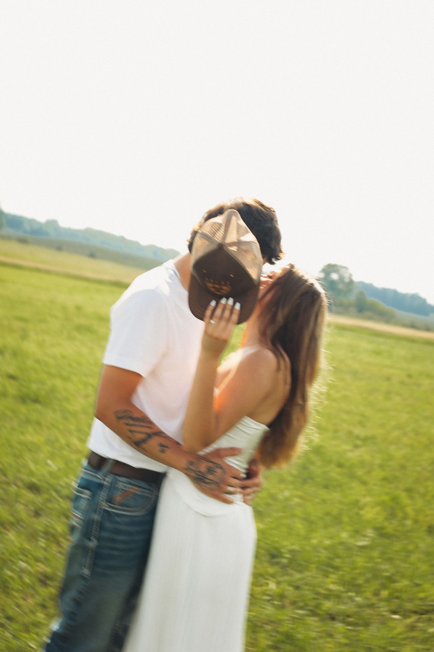 Couple kissing in a sunlit green field, man with tattoos in white tee and jeans, woman in white dress.