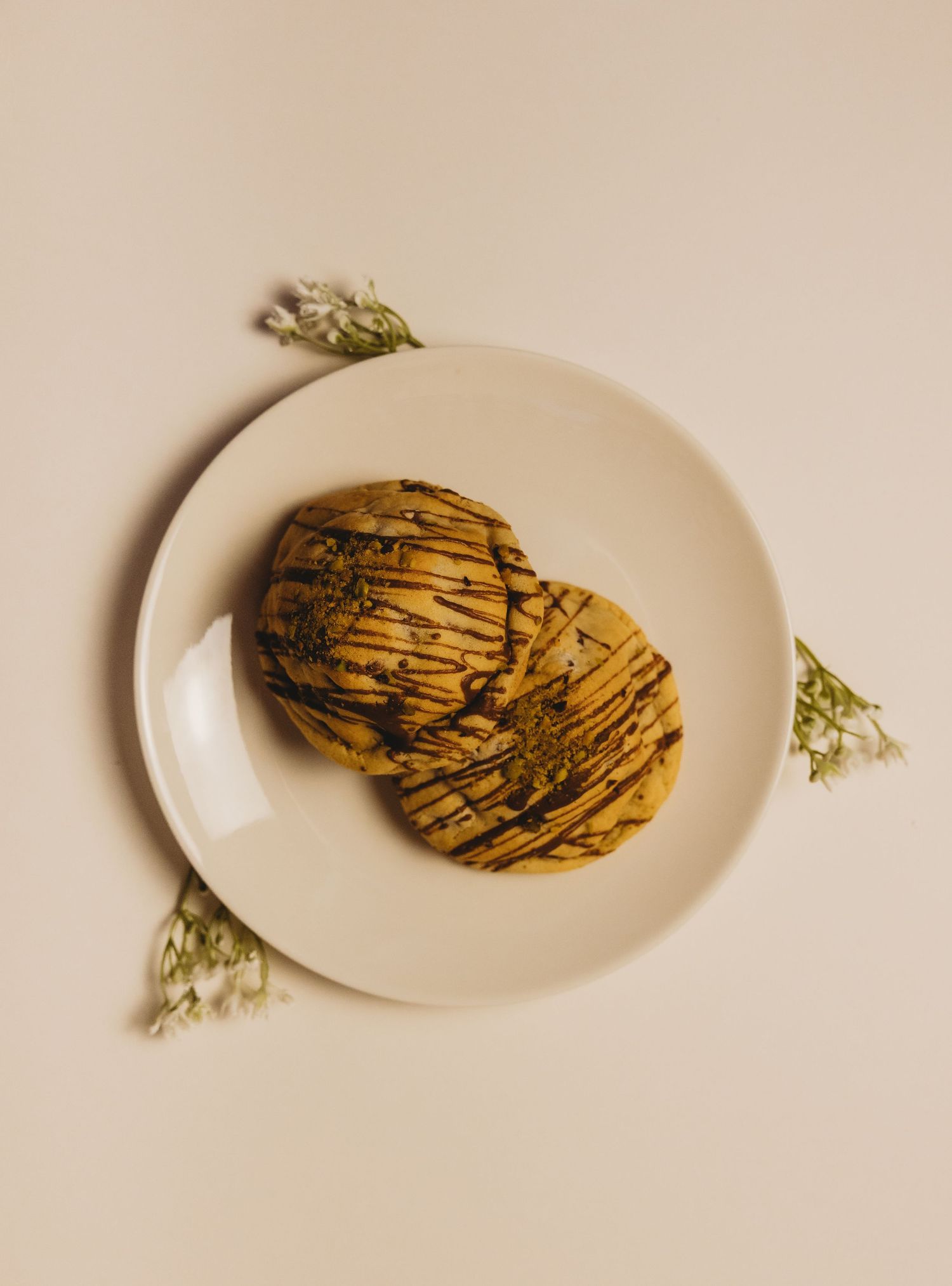 Two golden cookies drizzled with chocolate on a white plate, garnished with delicate white flowers.