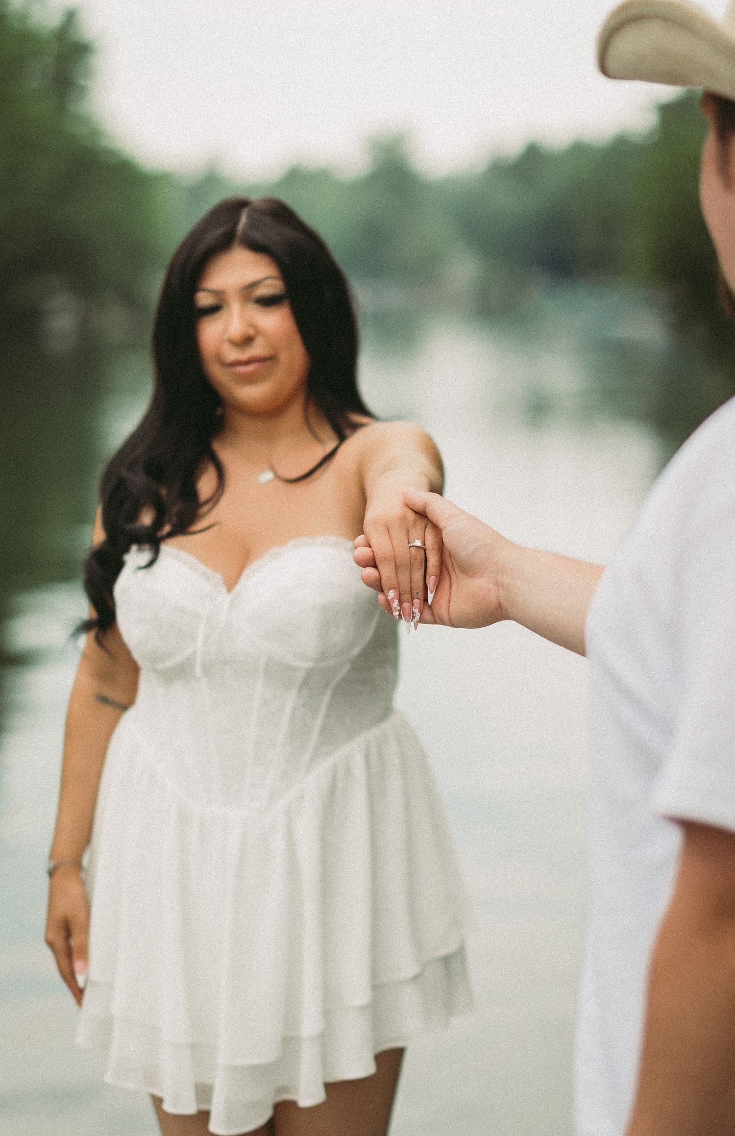 Woman in white summer dress poses by waterfront during engagement photoshoot.