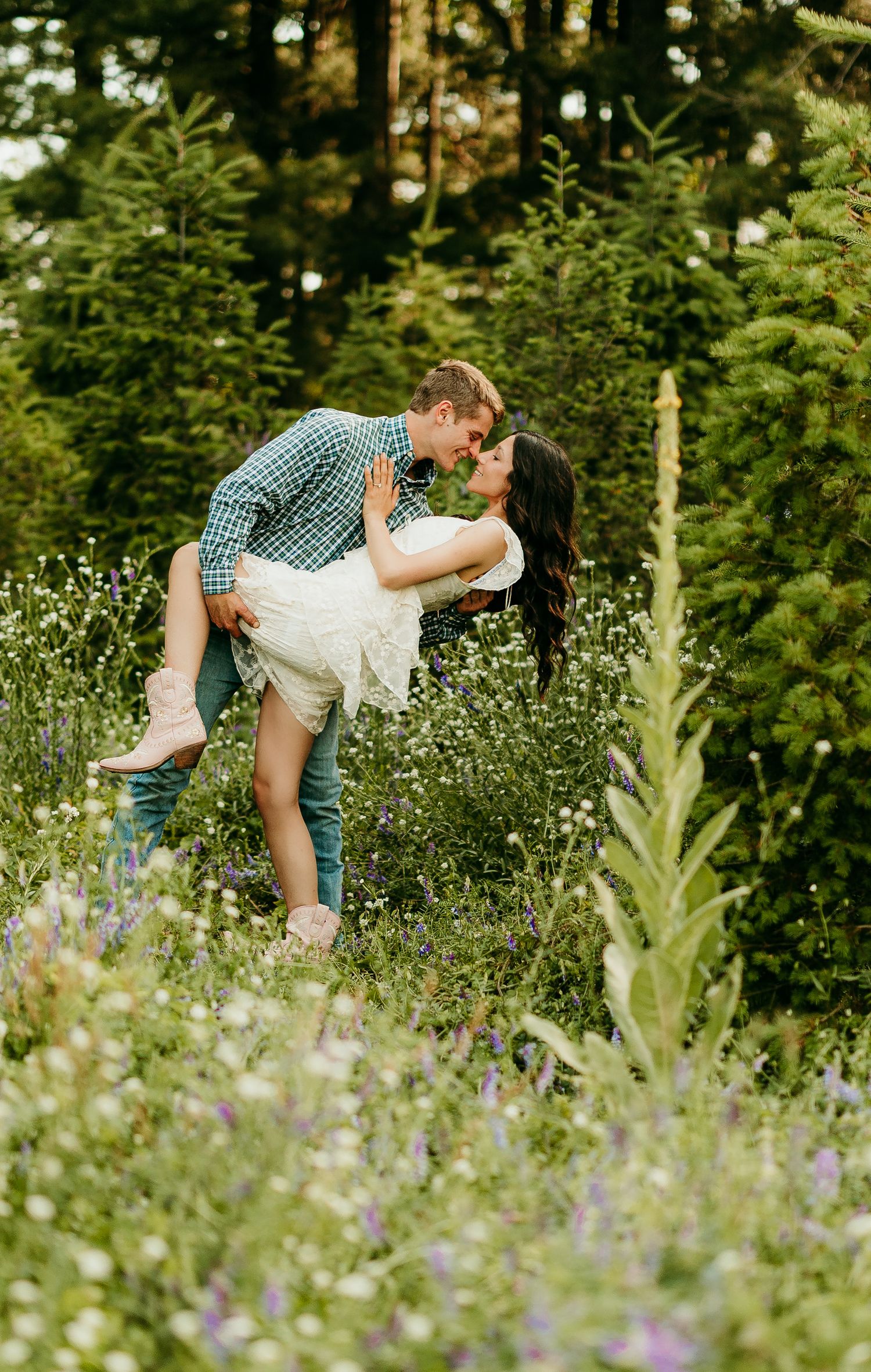 A couple embraces romantically in a lush meadow filled with wildflowers and evergreen trees on a sunny summer day.