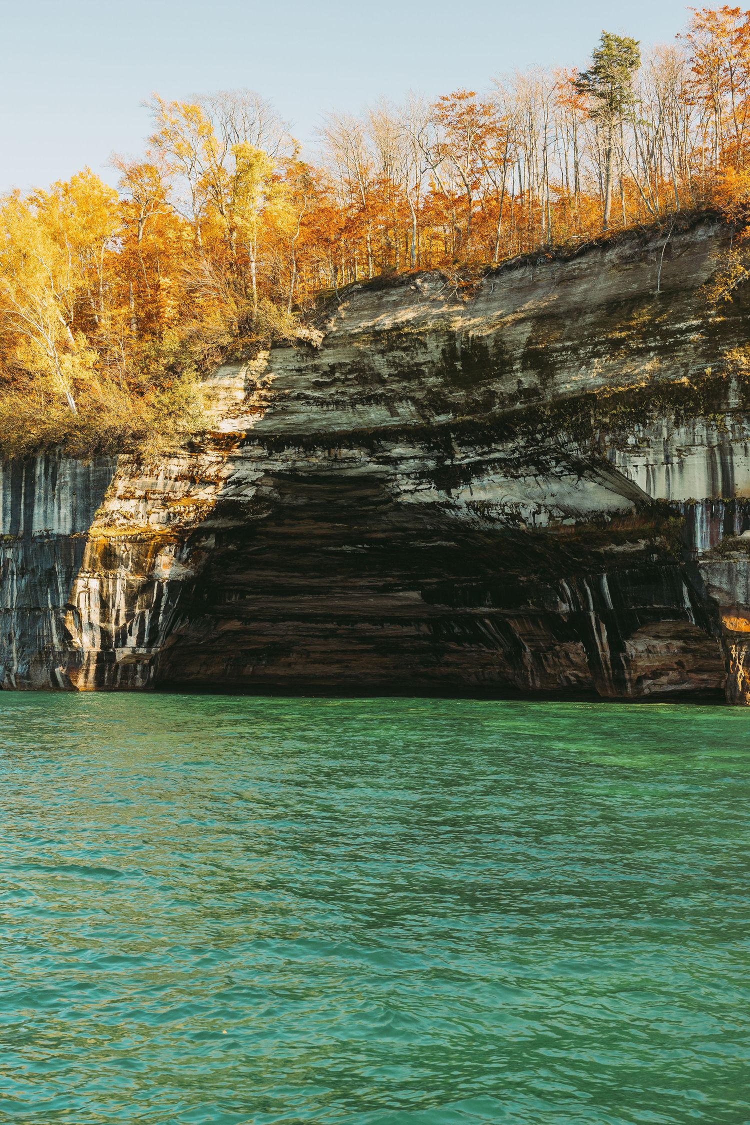 Layered rock formations tower over crystal clear waters with golden fall foliage on top.